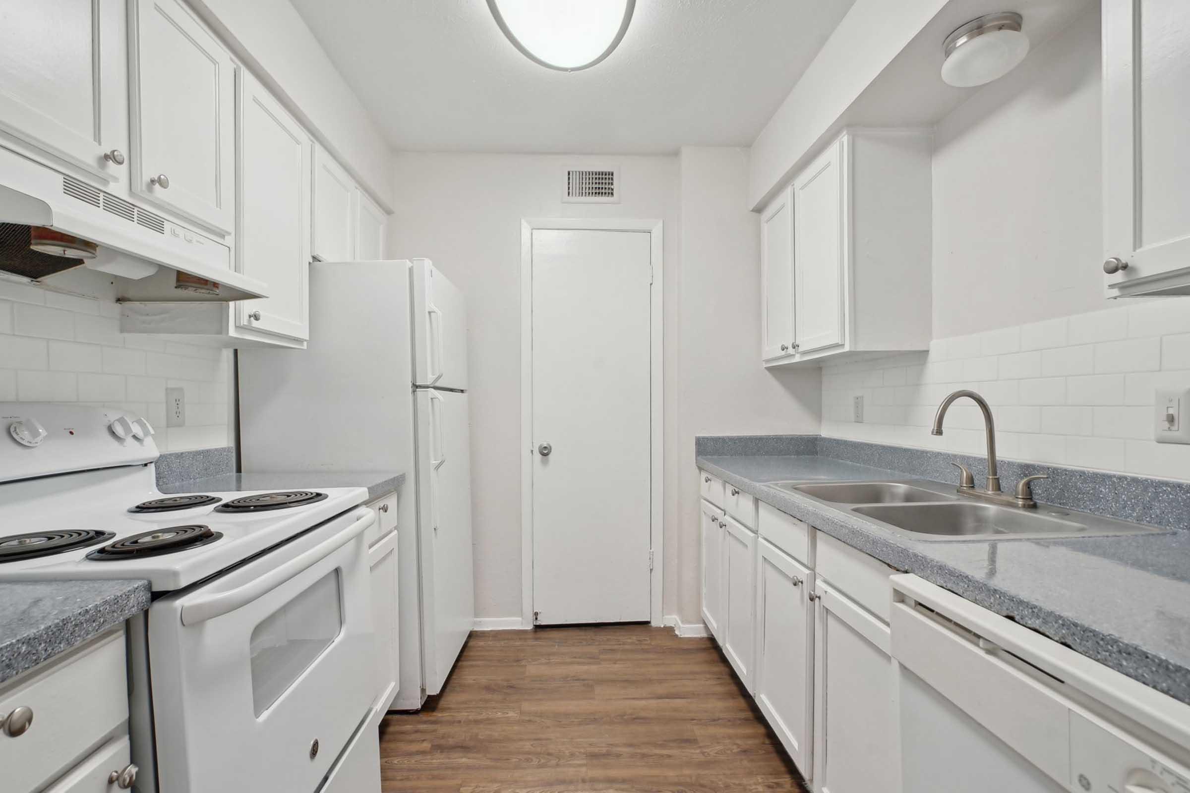 A clean, modern kitchen featuring white cabinetry, a white stove, and a double sink. The countertops are gray, and there are appliances including a refrigerator. The walls are light-colored, and there is a door leading to another space. The flooring is a wood-like design.