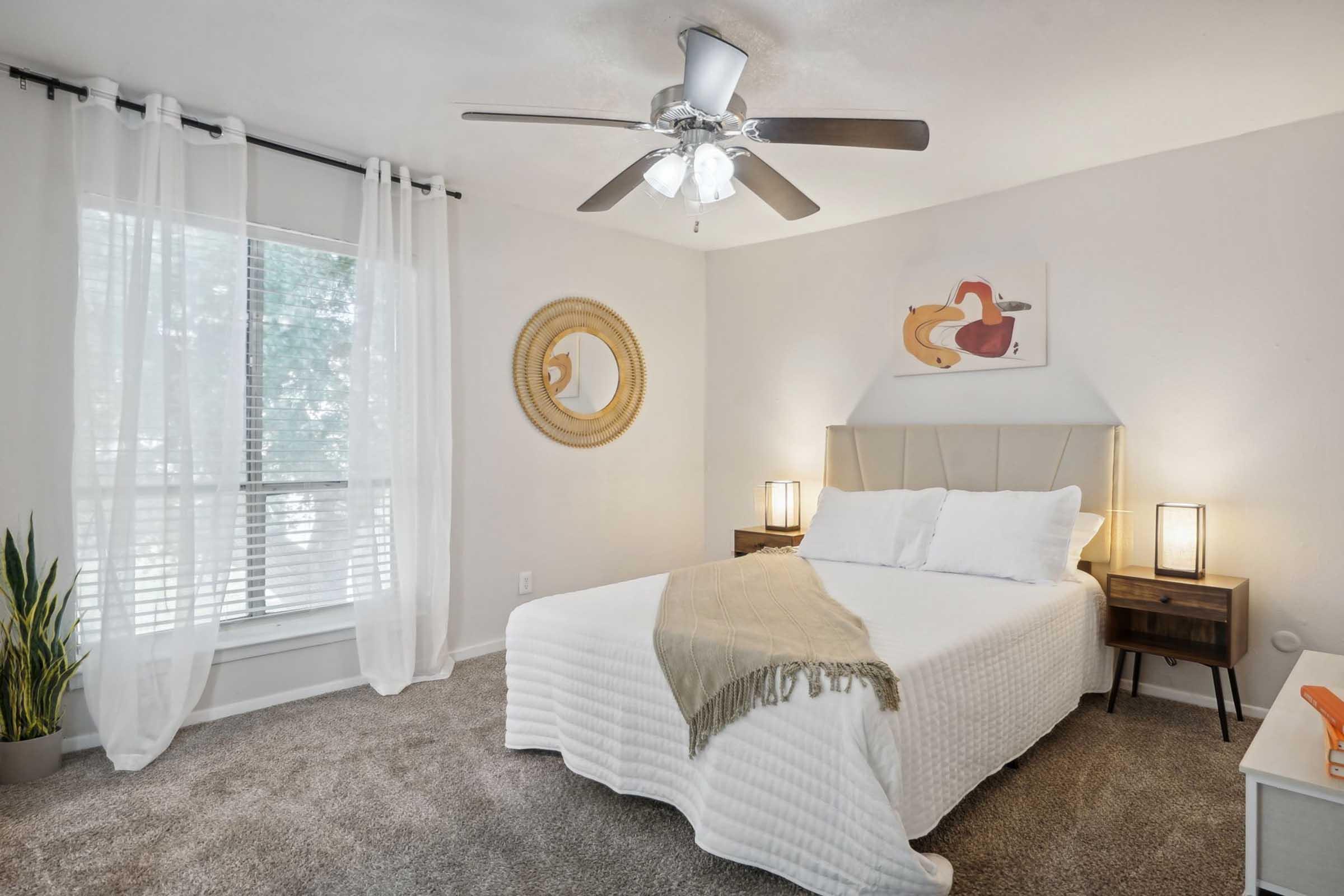 A cozy bedroom featuring a white bed with a textured blanket, two bedside tables with lamps, and a round mirror on the wall. A colorful artwork hangs above the bed. The room is well-lit by a ceiling fan and natural light streaming through sheer curtains, with a small plant in the corner.