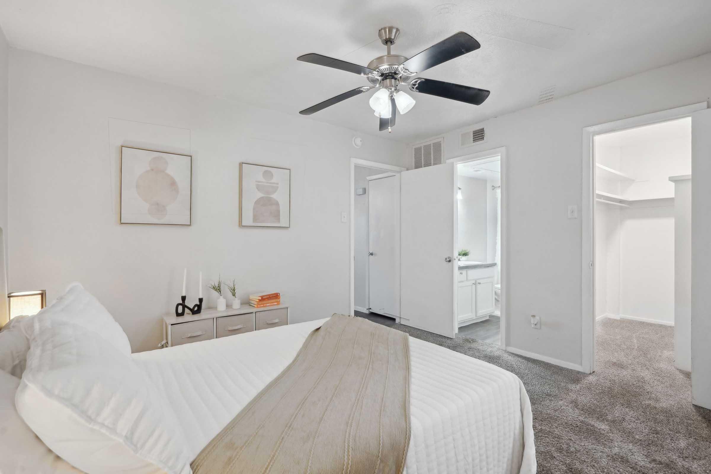 Bright and cozy bedroom featuring a ceiling fan, neutral-toned decor, and a comfortable bed with a textured blanket. Two framed artworks adorn the wall, and there’s a dresser with candles. A doorway leads to an adjoining room with a bathroom and a walk-in closet, all complementing the serene atmosphere.