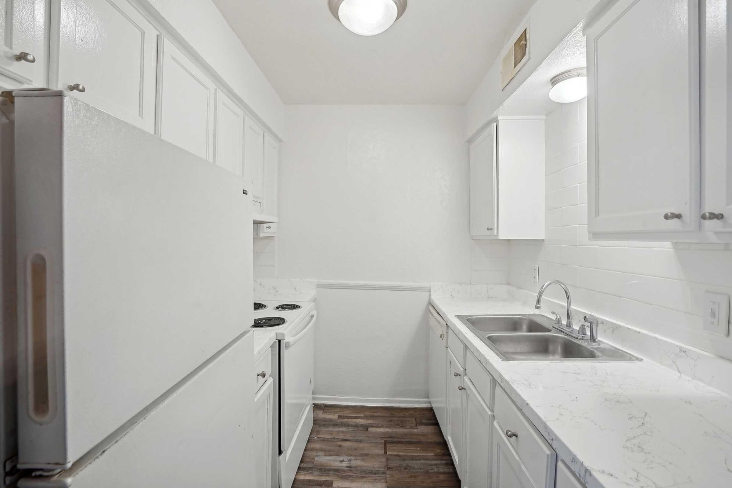 A modern, small kitchen featuring white cabinetry, a double sink, and a sleek marble countertop. The kitchen is equipped with a refrigerator and an electric stove. Bright lighting illuminates the space, highlighting the clean, minimalist design and wooden floor.