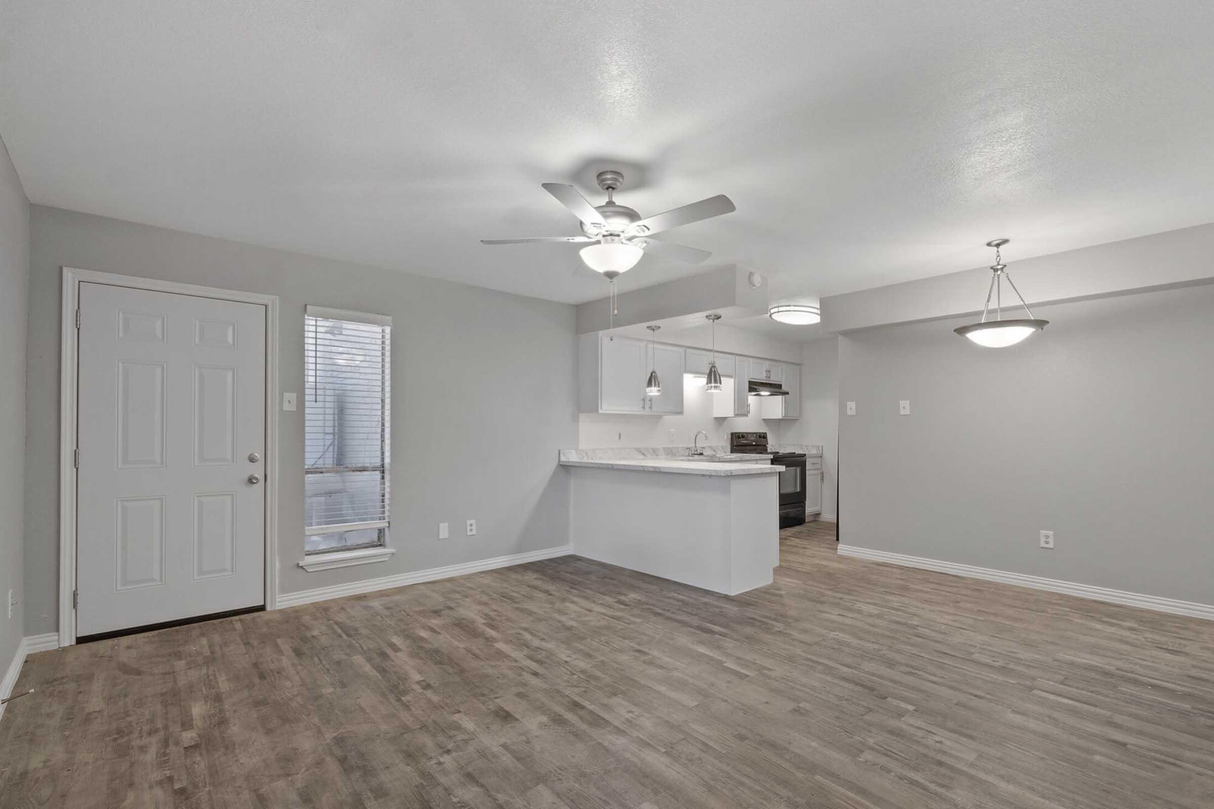 Modern interior of an apartment showcasing an open-concept living space. Features include light gray walls, a ceiling fan, a stylish kitchen area with pendant lighting, and large windows allowing natural light. The flooring is a warm wood tone, creating a cozy atmosphere.