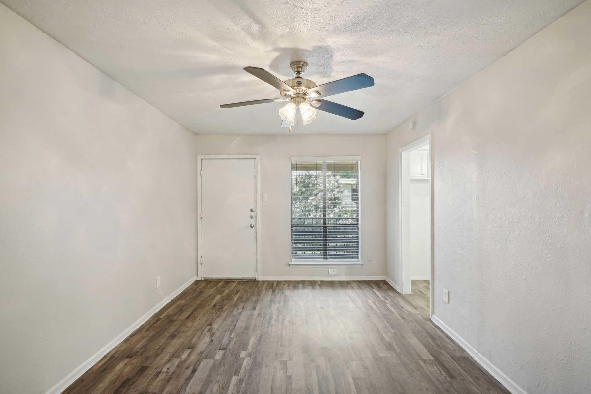 A spacious, light-colored living room featuring a ceiling fan with five blades, a front door on the left, a window with horizontal blinds, and a view of a small balcony. The flooring is wood-like laminate, and the walls are painted in a neutral tone, creating a welcoming ambiance.
