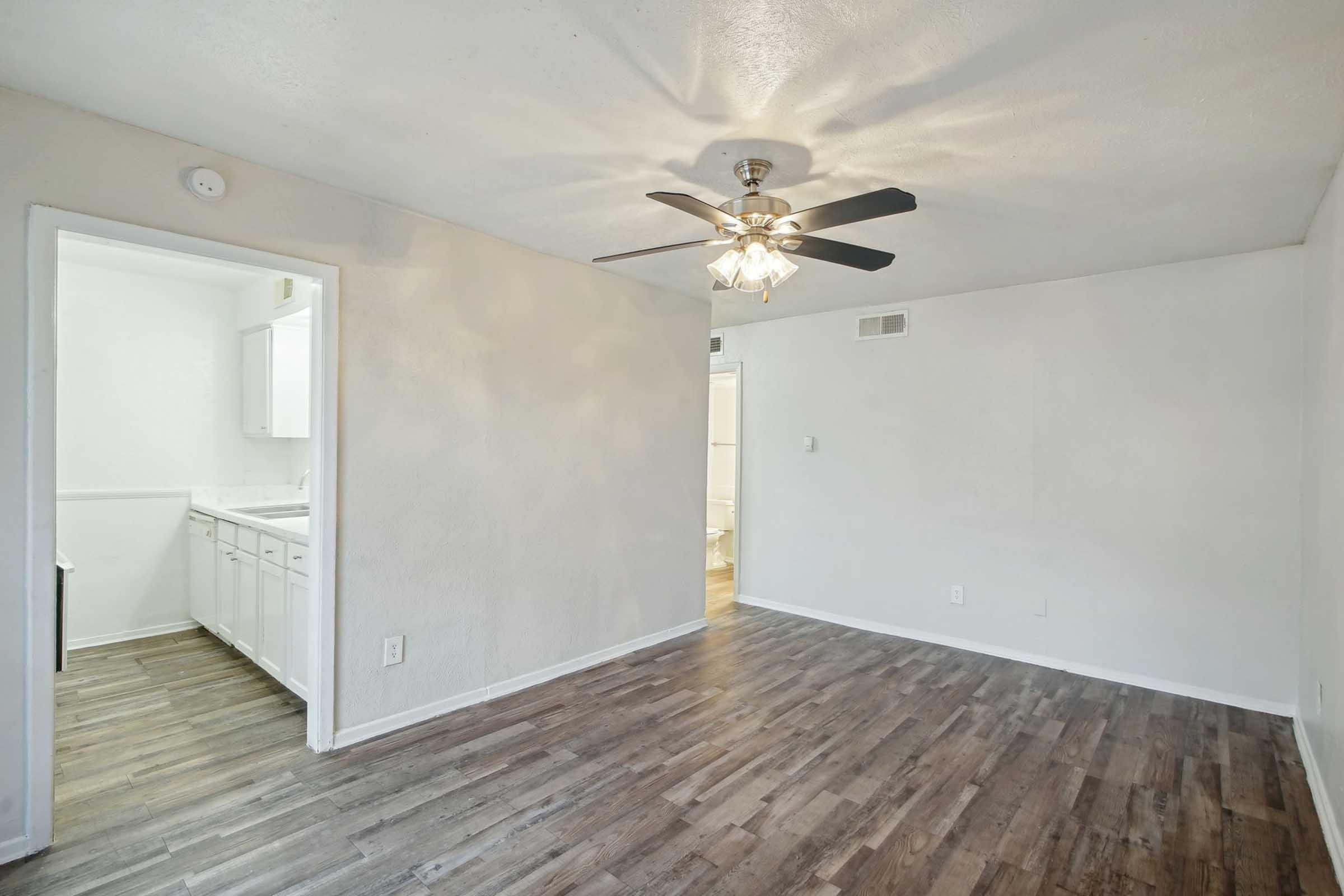 A spacious living area with light-colored walls and a ceiling fan. The flooring is wooden, and there is a doorway leading to a kitchen area. Natural light is present, creating a warm atmosphere. The overall design is modern and minimalistic, making it suitable for various interior styles.