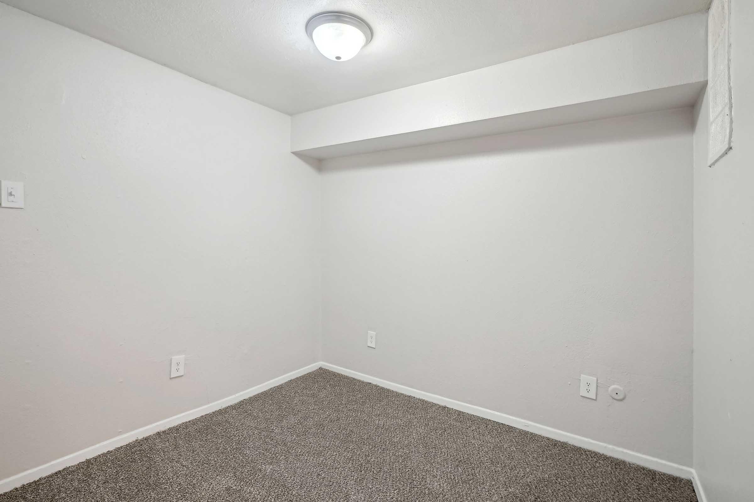 Empty room with light-colored walls and a ceiling light fixture. The floor is covered in carpet, and there are no furniture or decorations. One wall has a small section that visibly extends inward.