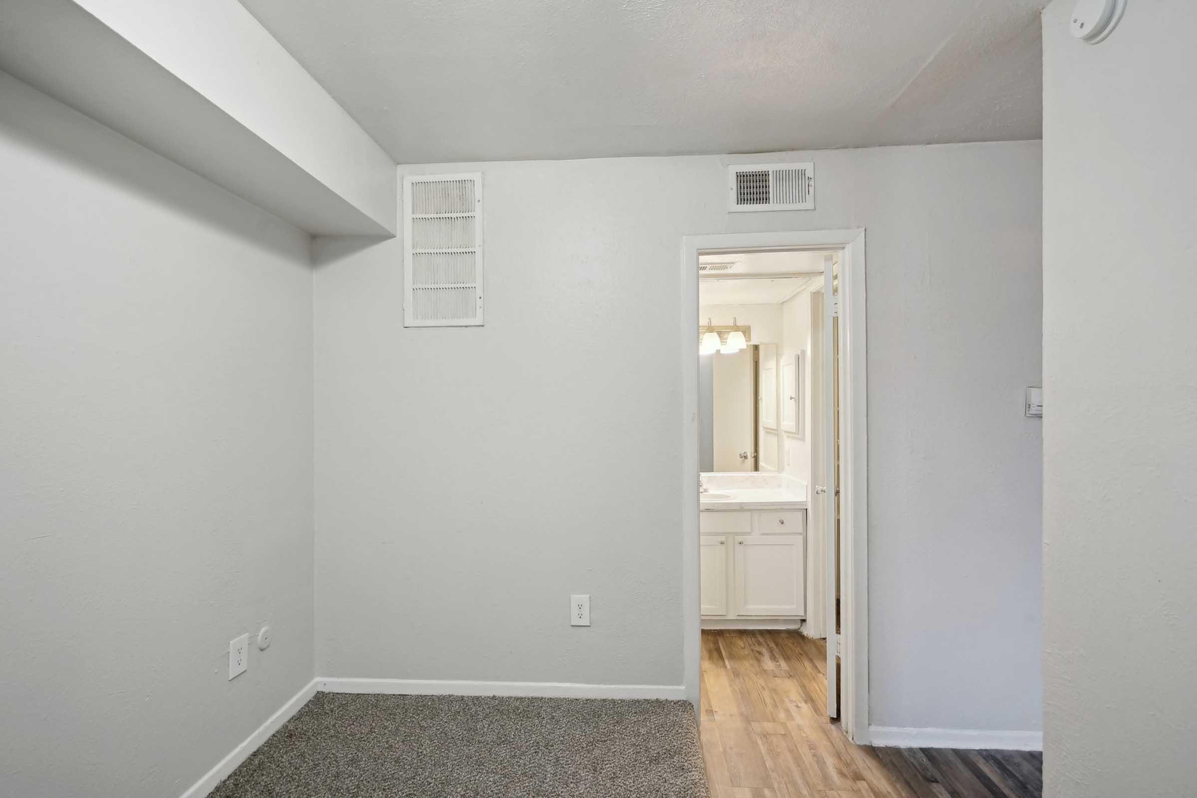 A plain, light-colored room featuring textured walls and carpeted flooring. There is a doorway on the right leading to a bathroom area with white cabinetry and a mirror. The room has minimal furnishings and natural light, creating a simple, functional space.