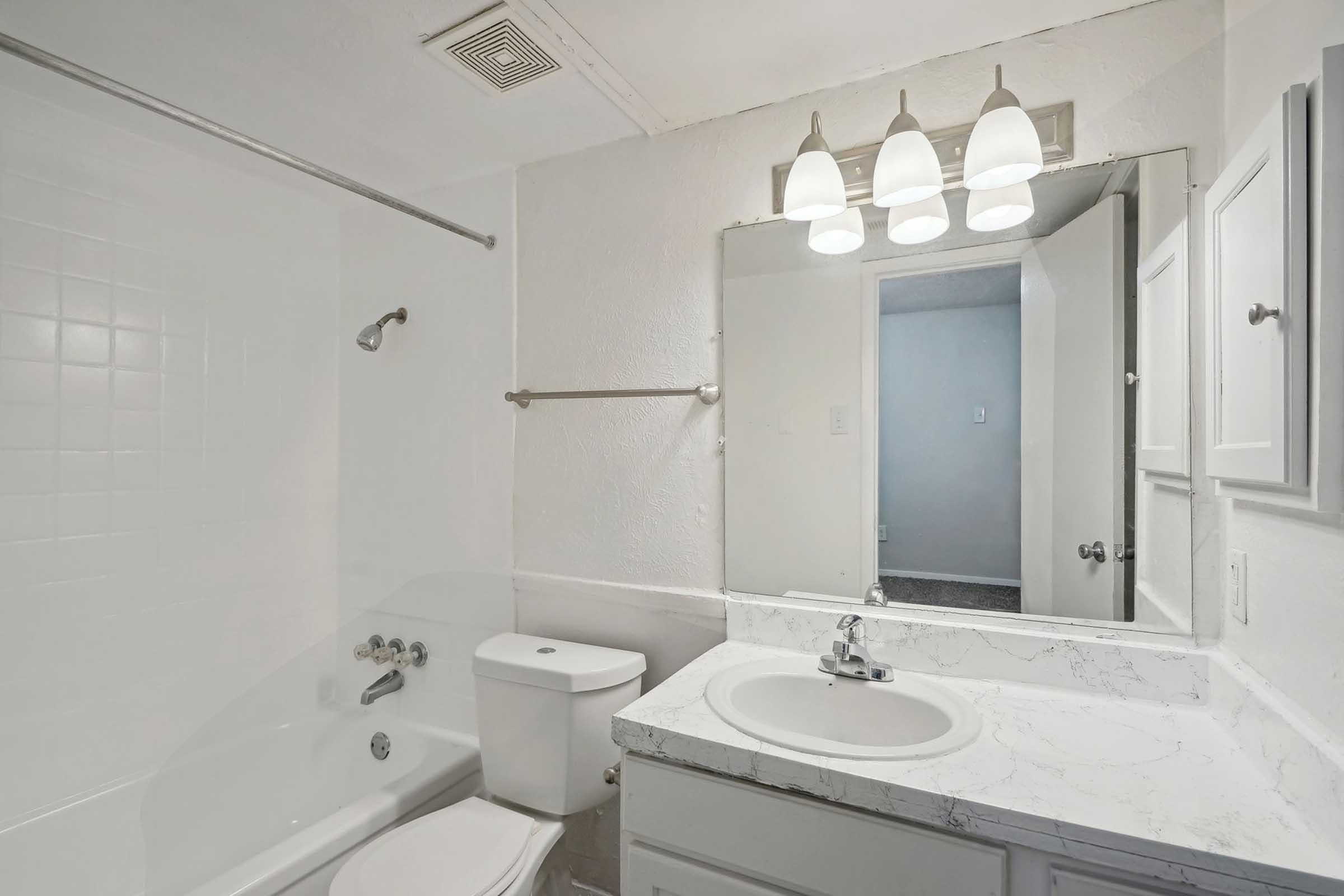 A clean, modern bathroom featuring a bathtub, toilet, and a vanity with a sink. The white walls and light-colored countertops enhance the bright atmosphere, while a mirror above the sink reflects the light from three pendant fixtures. A door leads to a separate room in the background.