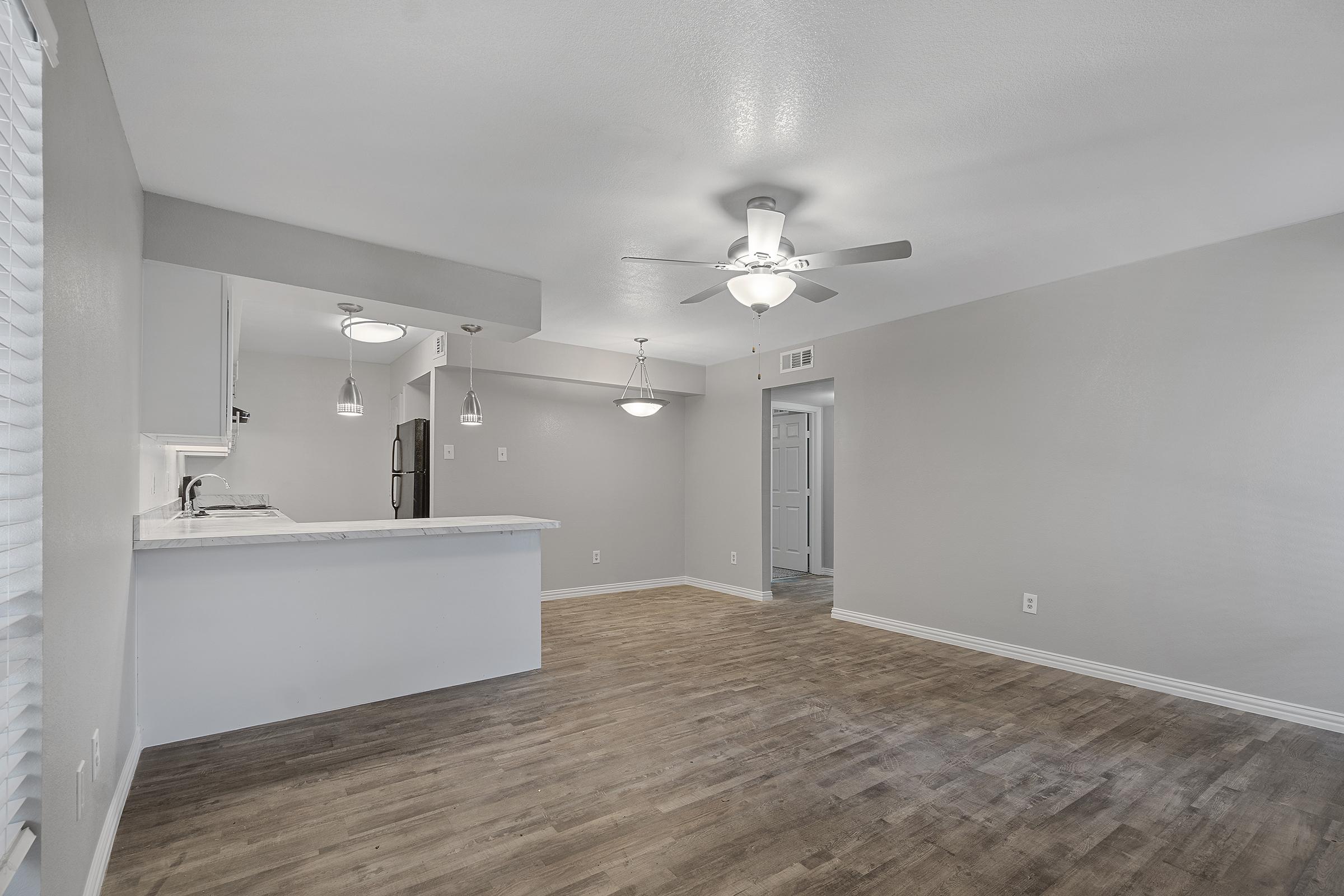 A modern, open-concept living space featuring a light gray color scheme. The room includes a ceiling fan, pendant lighting, and a kitchen area with a countertop. There are large windows allowing natural light, and the flooring is a warm wood tone, creating a welcoming atmosphere.
