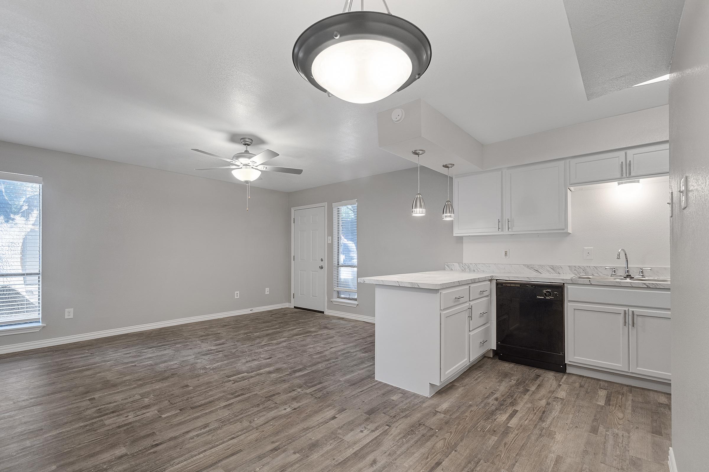 A spacious, modern kitchen and living area with light-colored walls, wood-like flooring, and ample natural light. There is a ceiling fan, white cabinets, a marble countertop, a black dishwasher, and large windows. The layout is open and inviting, perfect for a contemporary home.
