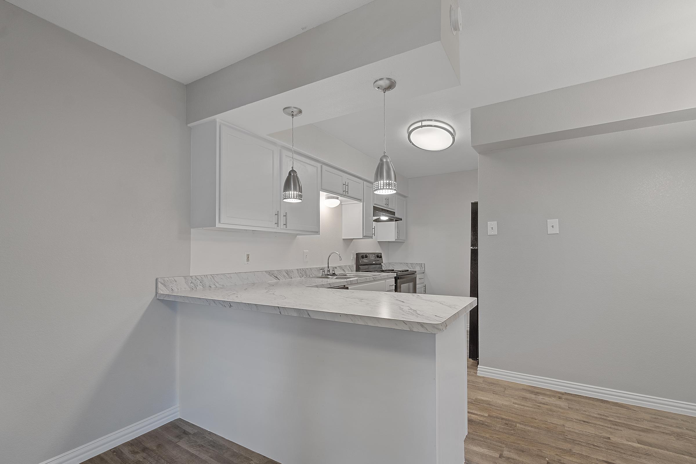A modern kitchen with a marble countertop and stainless steel appliances. There are two pendant lights hanging above the counter. The walls are painted in a light gray color, and the floor is a light wood laminate. The space appears clean and bright, suitable for both cooking and entertaining.