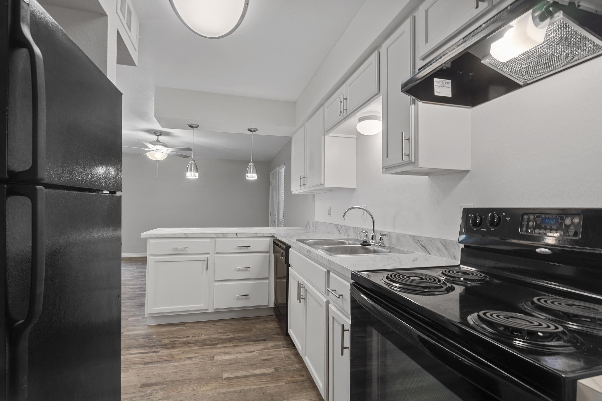 Modern kitchen featuring a black refrigerator and black stove, white cabinetry, and a marble countertop. The space is well-lit with pendant lights and has wood flooring, creating a bright and inviting atmosphere. The layout includes a sink and an island area for additional prep space.