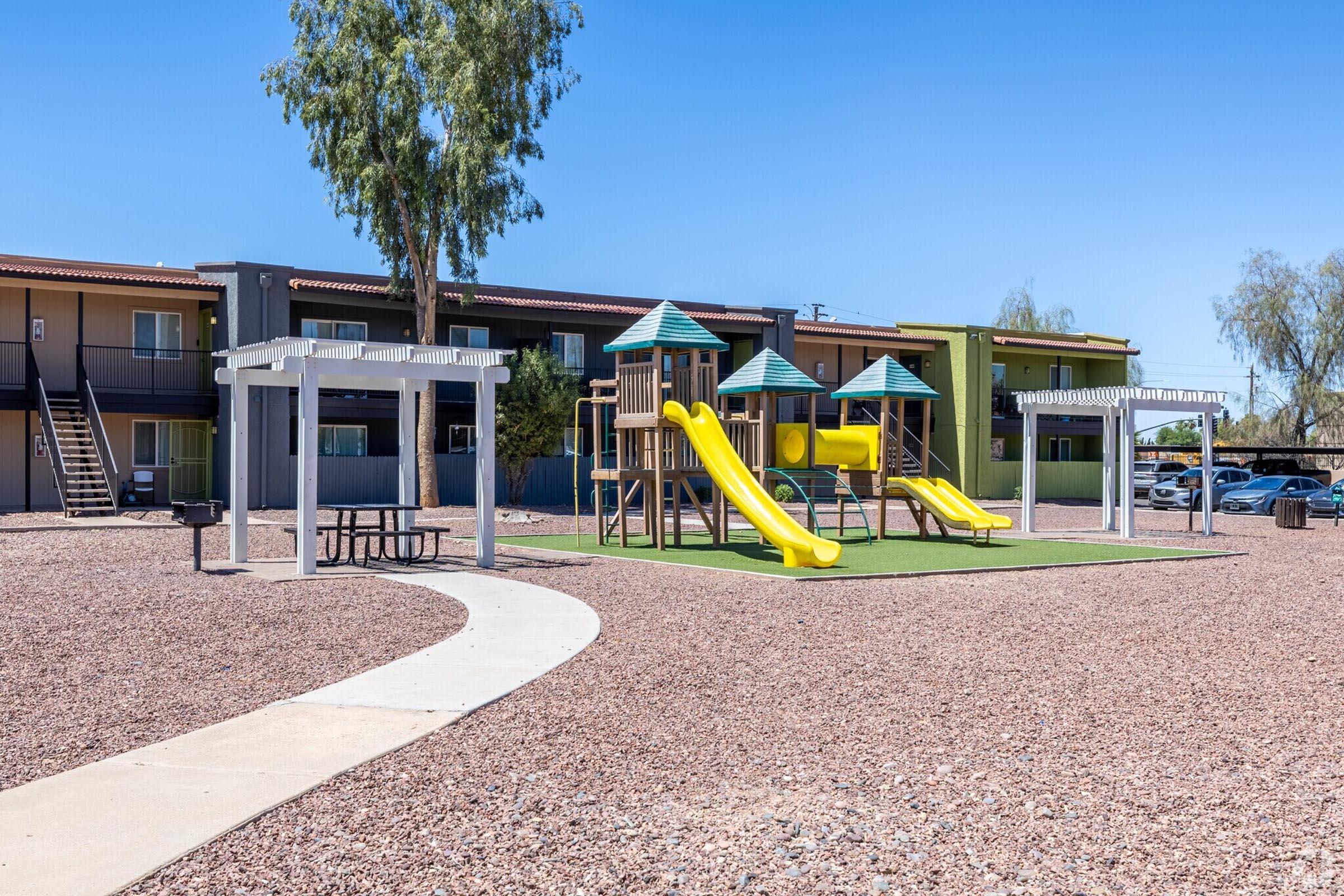 A playground featuring yellow slides and a wooden play structure is set on a grassy area, surrounded by a bed of gravel. In the background, there are low-rise apartment buildings with balconies and a clear blue sky above. A concrete path leads through the play area, with picnic tables visible nearby.
