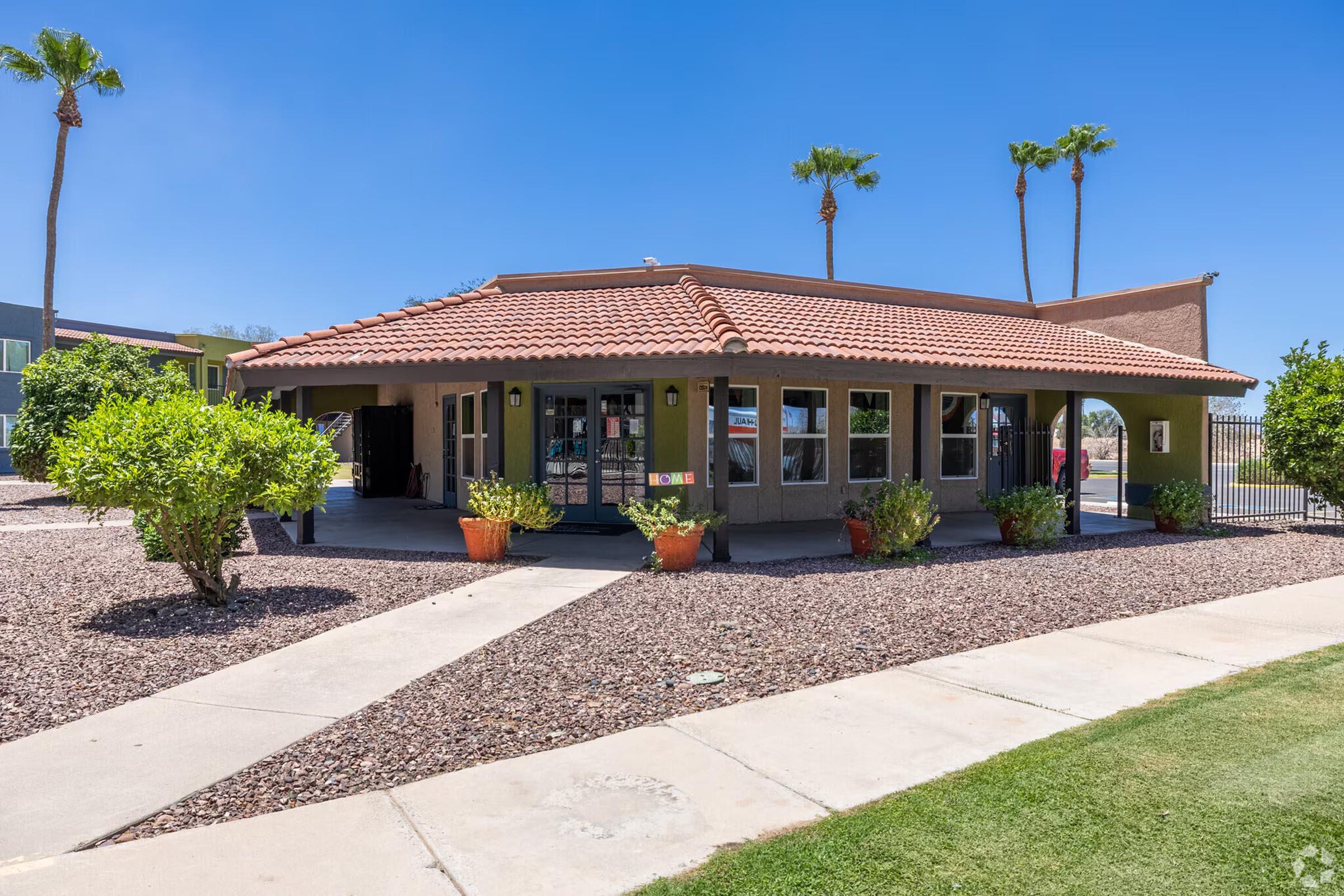 A single-story building with a terracotta tile roof, surrounded by landscaped areas featuring shrubs and small trees. There is a pathway leading to the entrance, and the building has large windows, suggesting a welcoming atmosphere. Palm trees are visible against a clear blue sky.