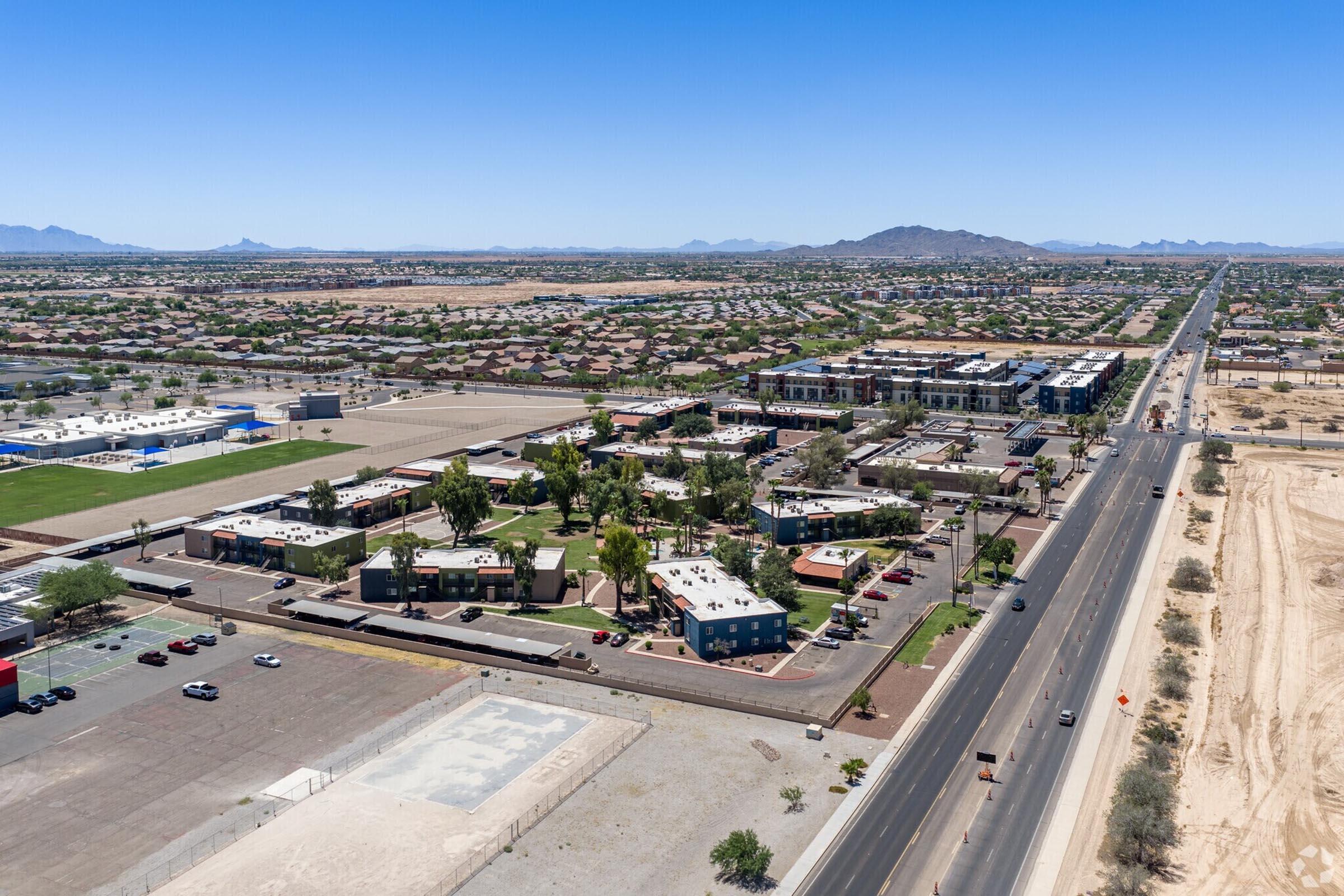 Aerial view of a suburban area featuring a residential neighborhood, green parks, and a nearby road. The landscape includes distant mountains and a clear blue sky. Parking lots are visible, along with a mix of buildings and open spaces. In the foreground, an empty lot is seen, suggesting potential development.