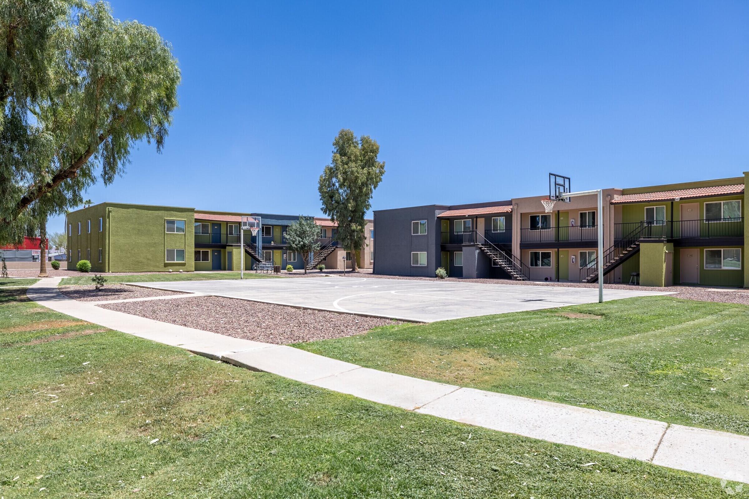 A wide view of a residential complex featuring two-story apartment buildings. The buildings are positioned around a central outdoor area with a basketball hoop. Lush green grass and trees surround the courtyard, under a clear blue sky.