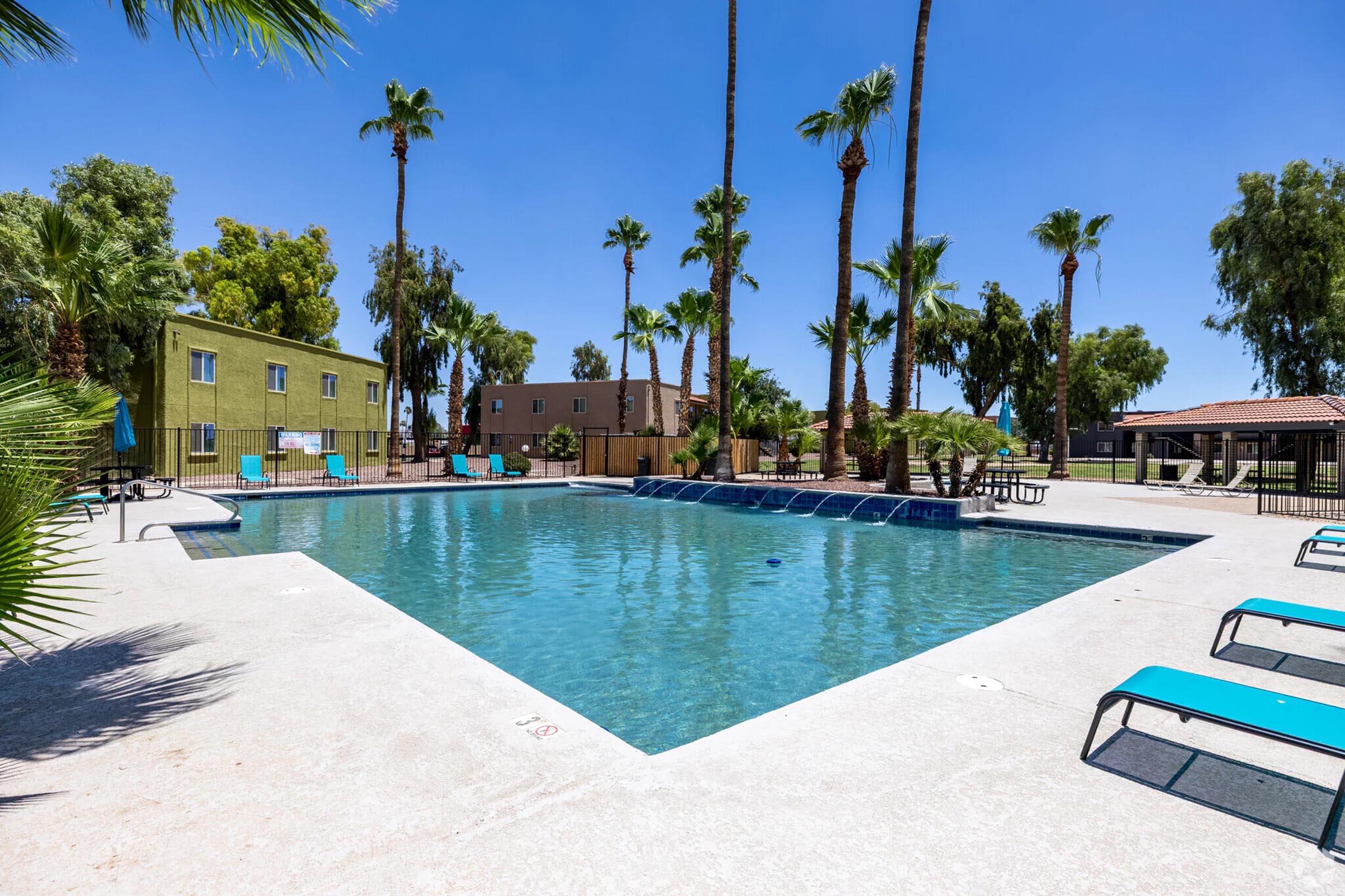 A clear blue swimming pool surrounded by palm trees and lounge chairs. The area is sunny with a few residential buildings in the background, providing a relaxing atmosphere ideal for leisure and recreation. The pool features a shallow entry and is fenced for safety.