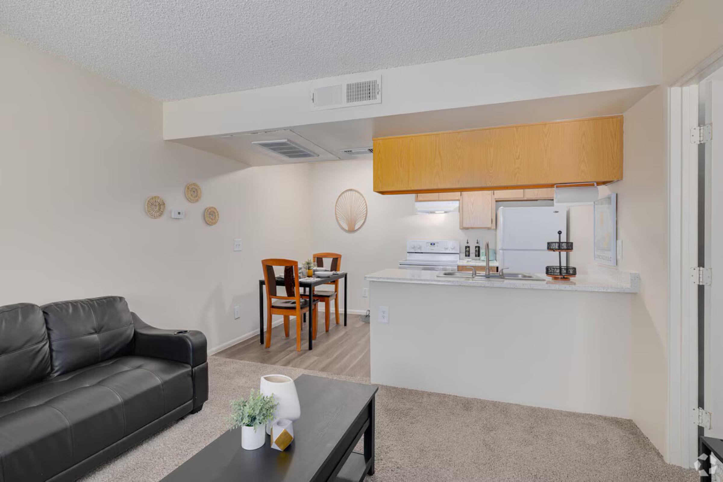 A cozy living space featuring a black sofa, a coffee table, and a small dining area with wooden chairs. The kitchenette has wood cabinets, a white stove, and a refrigerator. The room has light-colored walls and a carpeted floor, with decorative wall elements adding a touch of style.