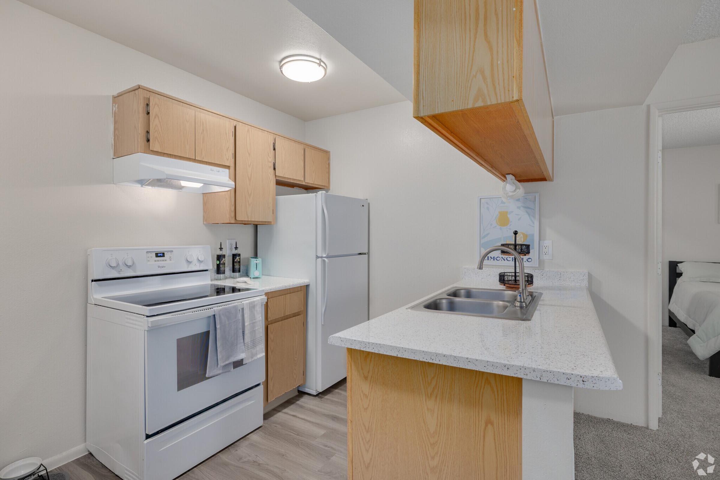A modern kitchen featuring light-colored cabinets, a white stove with an overhead microwave, a white refrigerator, and a countertop with a sink. The flooring is laminate, and there's a small bar separating the kitchen from the adjacent living area. Bright lighting enhances the overall space.