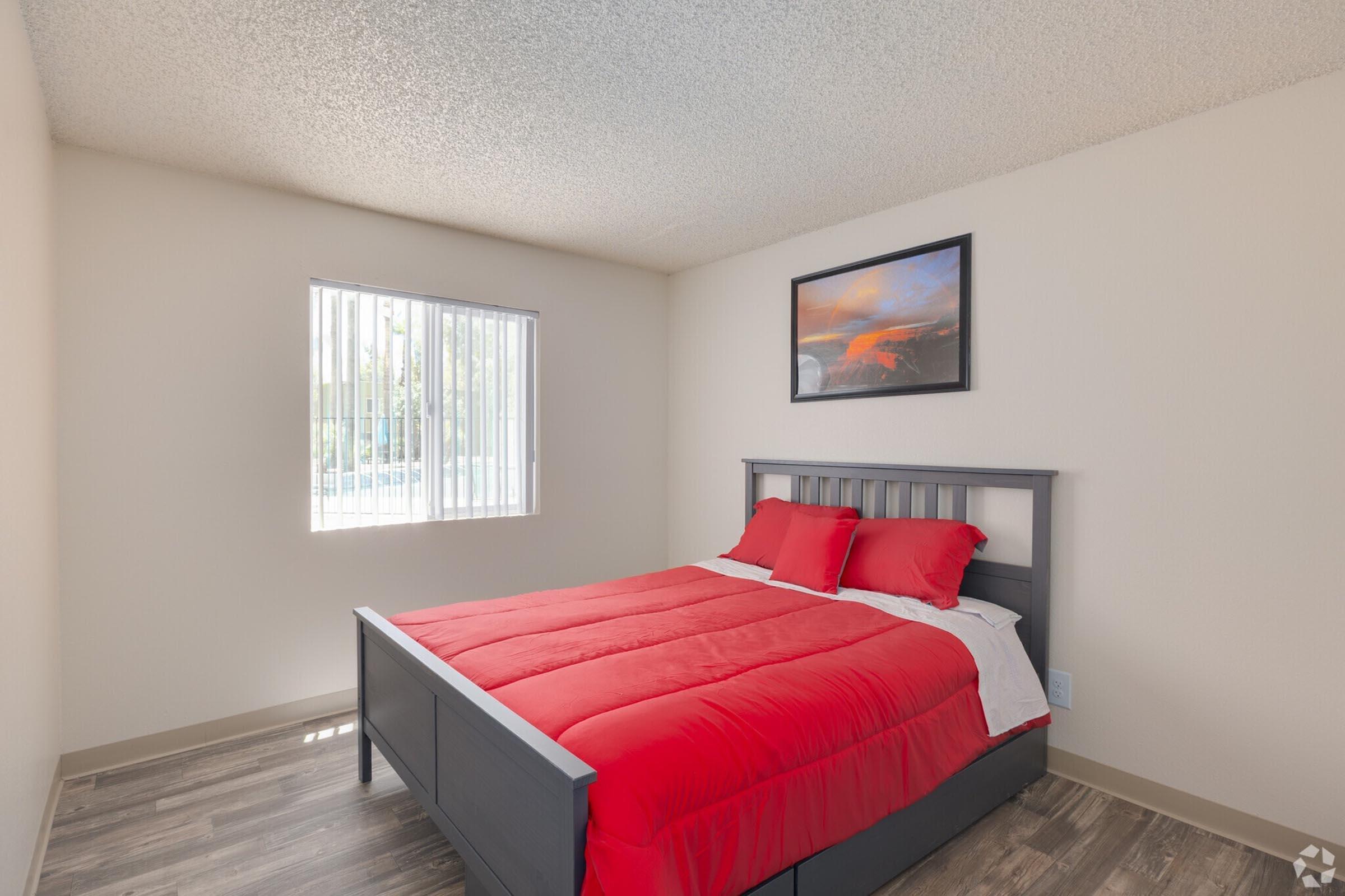 A bright, clean bedroom featuring a gray wooden bed with a red comforter and matching pillows. There's a window with blinds letting in natural light, and a scenic landscape painting hangs on the wall above the bed. The flooring is a light-colored laminate, giving the room a modern feel.