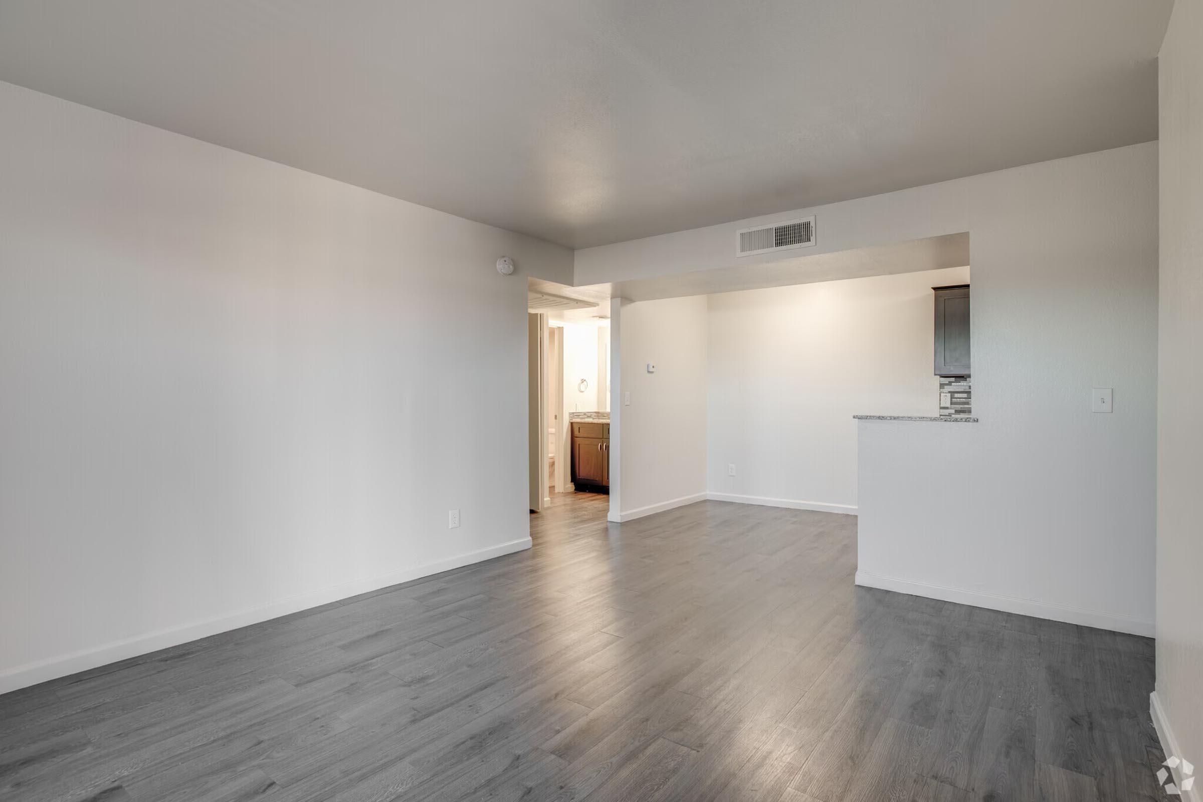 A spacious, empty living area with light-colored walls and wooden flooring. There is an open layout leading to a modern kitchen area in the background. A doorway to a bathroom is visible on the left, and the room is well-lit with natural light coming from an unseen window.