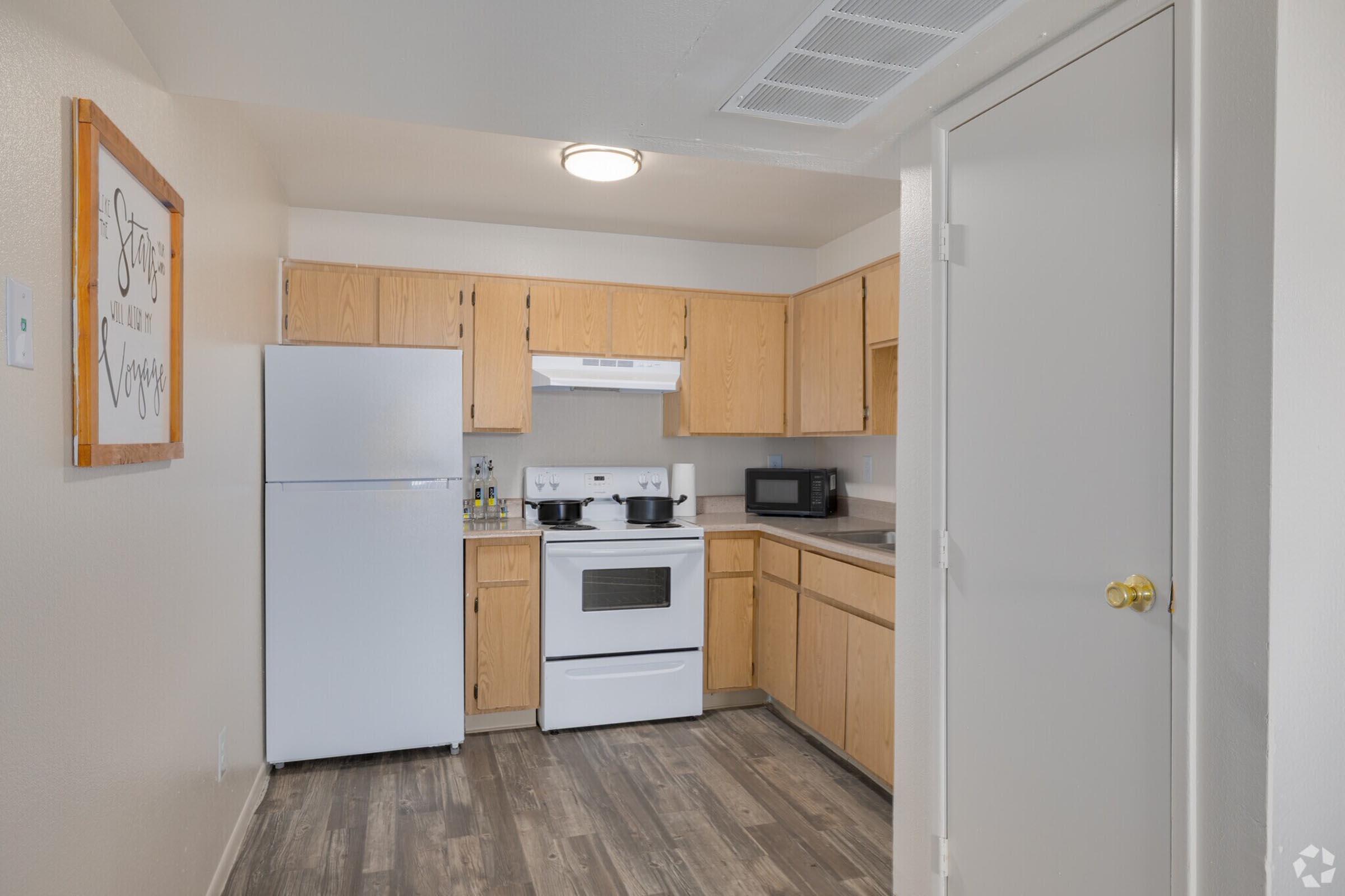 A clean, modern kitchen featuring wooden cabinets, white appliances including a refrigerator, stove, and microwave, along with a countertop. The flooring is a light wood laminate. A framed sign on the wall reads "Stay awhile." The space is well-lit, creating a welcoming atmosphere.