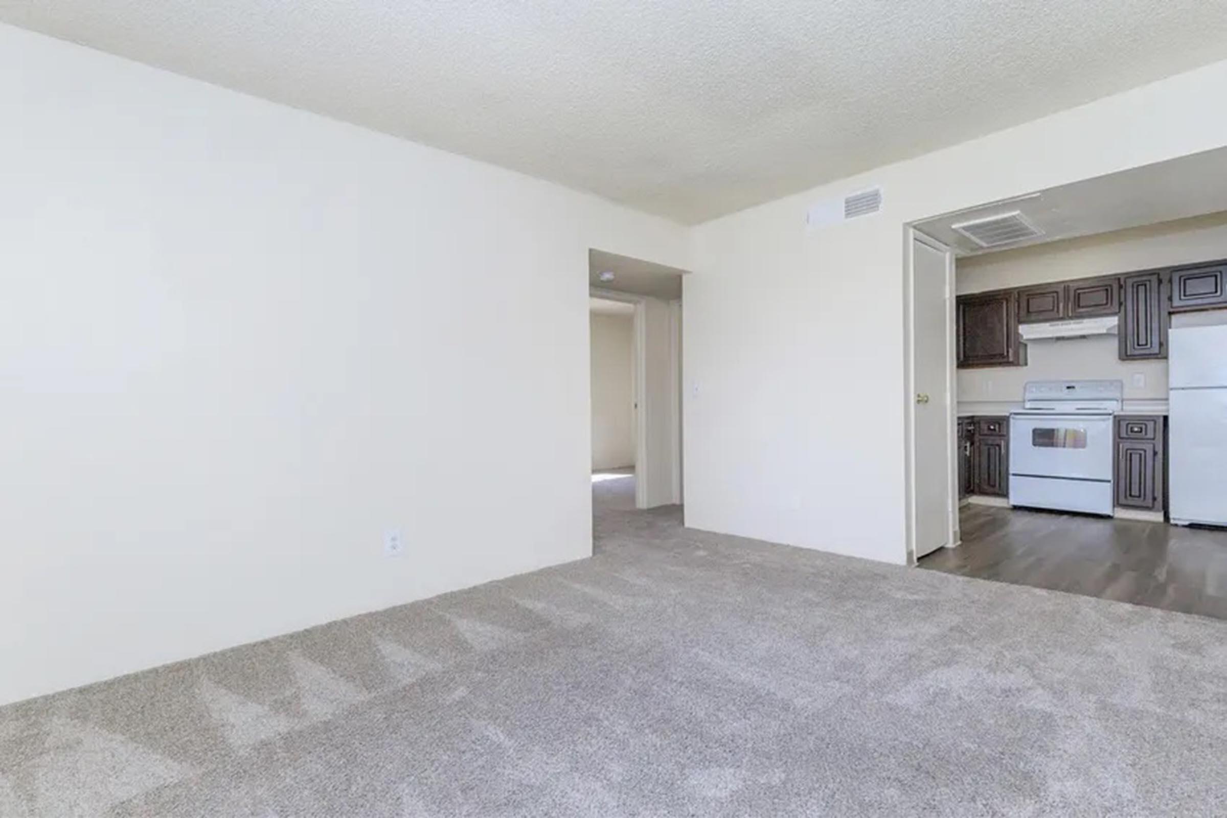 A spacious, light-colored living area featuring soft, plush carpeting. To the right, there's an open doorway leading into a kitchen with brown cabinetry and white appliances. The walls are painted in a neutral tone, enhancing the bright and airy feel of the space. Sunlight is visible from the adjacent room.