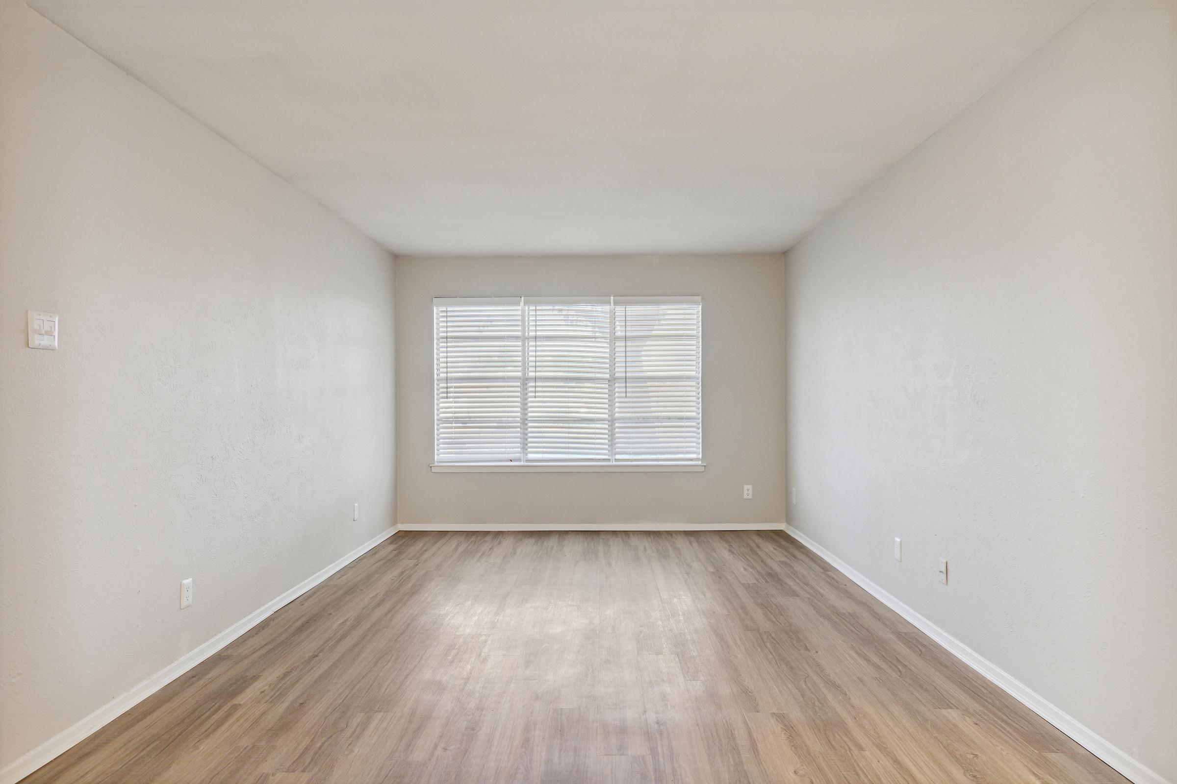 A spacious, empty room with light-colored walls and a wooden floor. There are large windows with white blinds letting in natural light, creating a bright, airy atmosphere. The room is free of furniture, emphasizing its openness and potential for various arrangements.