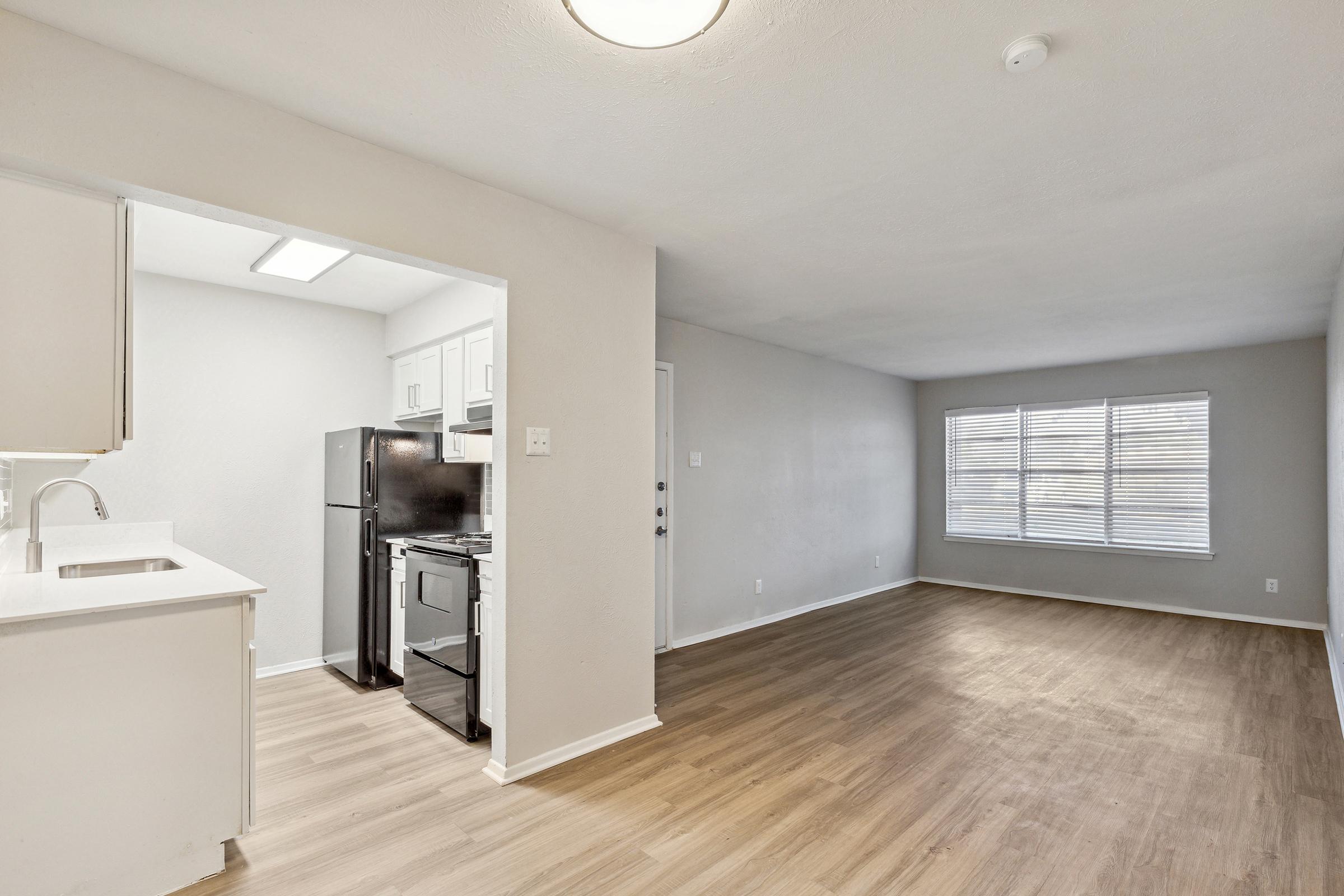 A spacious living area featuring light-colored walls and wood-like flooring. On one side, there is a kitchen with white cabinetry and stainless steel appliances, including a refrigerator and an oven. A large window allows natural light to fill the room, enhancing the open atmosphere.