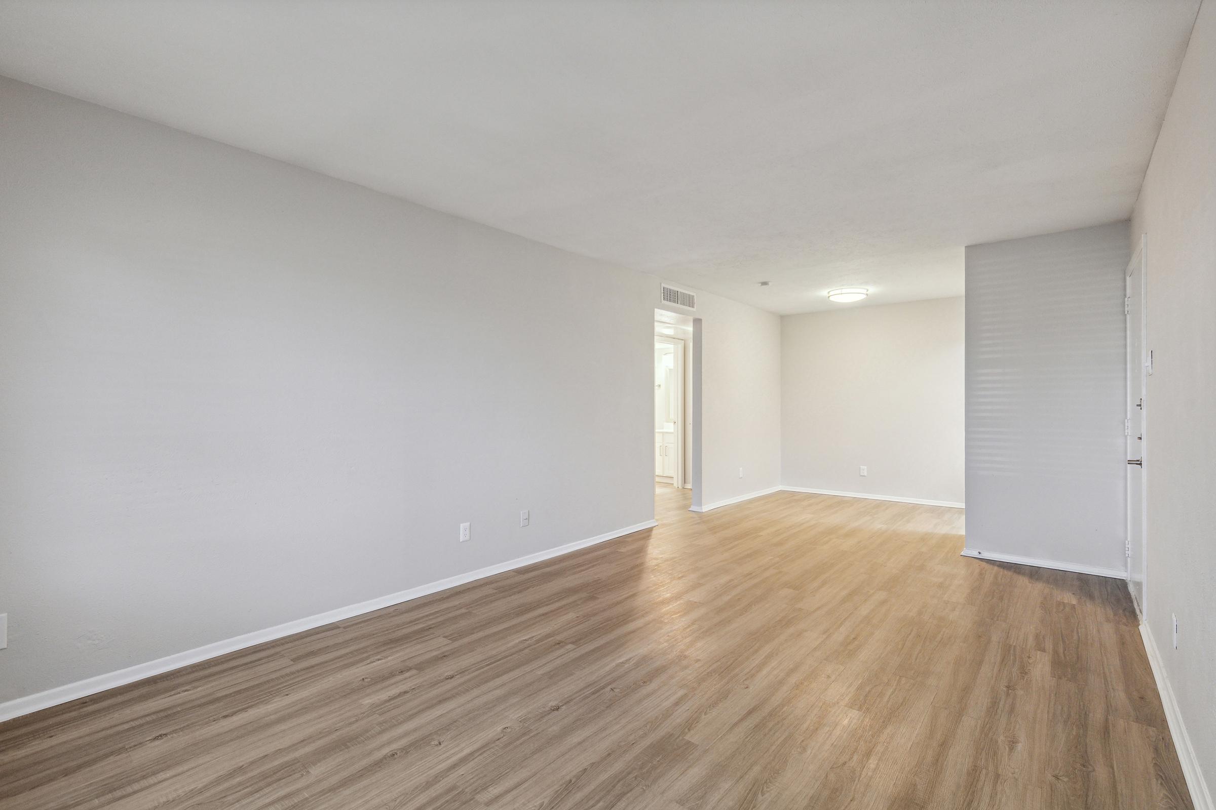 A spacious, unfurnished living room with light-colored walls and laminate flooring. Natural light fills the room from a nearby doorway, leading to a bathroom or kitchen area. The decor is minimal, creating a blank canvas for potential furnishings.