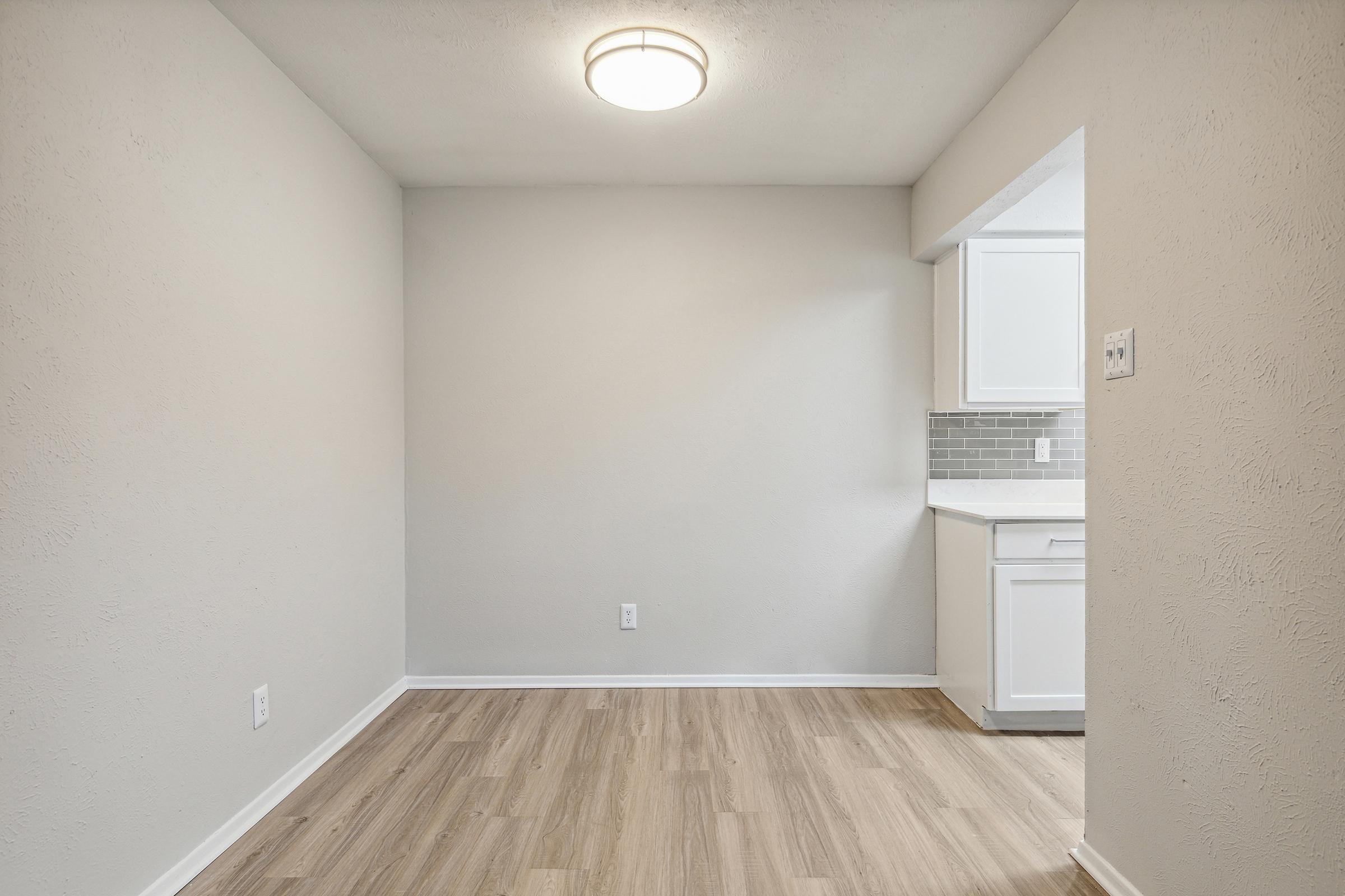A small, empty room with light gray walls and a wooden floor. A ceiling light fixture is present, and there is an open space leading to a kitchen area with white cabinets and a gray tiled backsplash. The overall ambiance is modern and minimalistic.