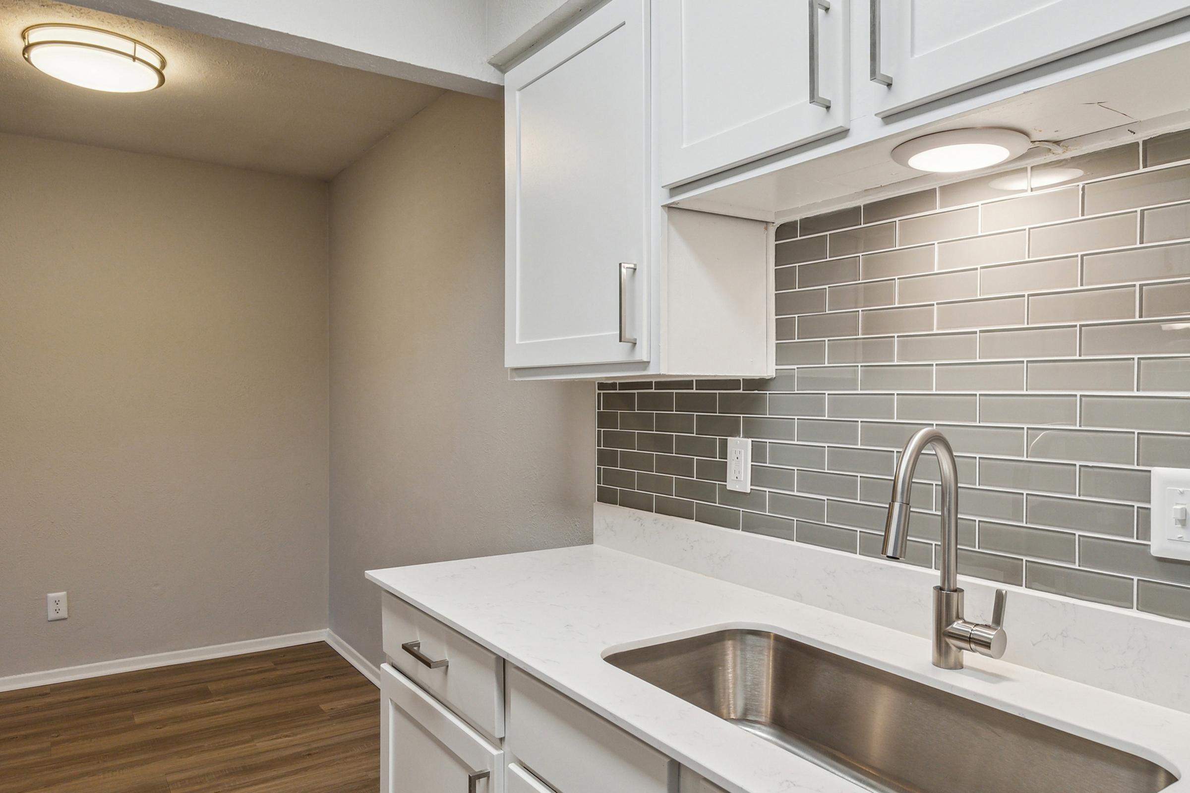 Modern kitchen with white cabinets and a stainless steel sink. The backsplash features grey tiles, and there is warm overhead lighting. A light-colored wall complements the overall design, while a wooden floor adds texture to the space.