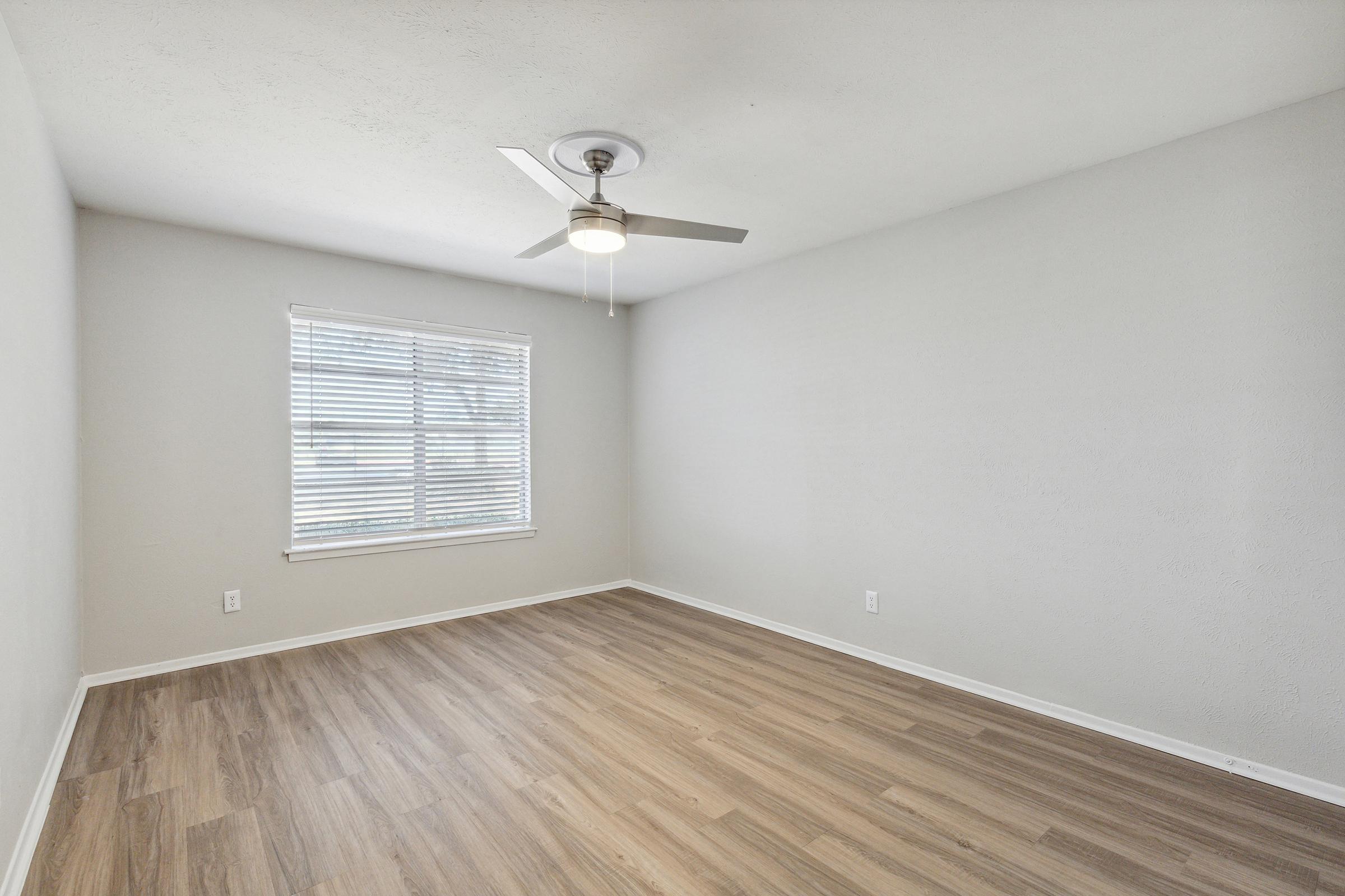 A spacious, empty room featuring light gray walls, a ceiling fan, and a large window with white blinds. The floor is covered in light-colored wood laminate, and there are no furnishings present, creating a bright and open atmosphere.