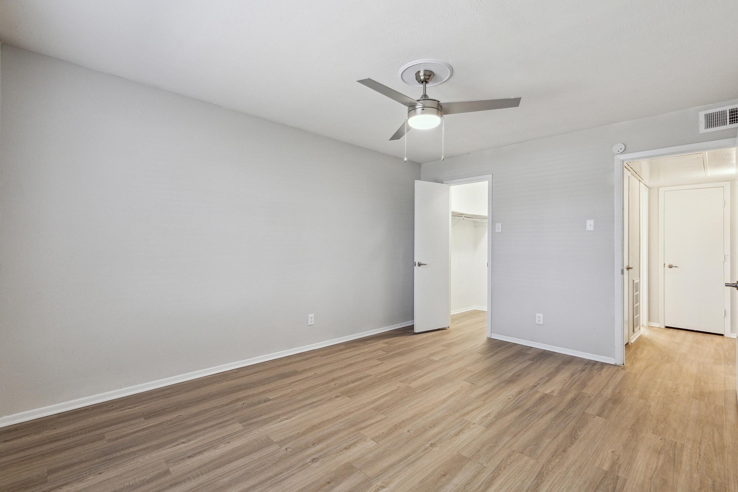 Empty room with light gray walls and wood flooring. A ceiling fan is installed in the center. There is a closet with sliding doors on the left side, and a doorway leading to another room is visible on the right. The space is bright and uncluttered, offering a minimalist aesthetic.