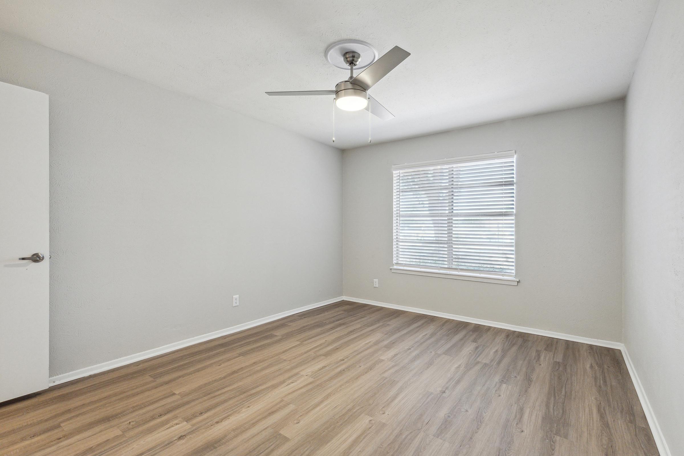 A well-lit, empty room featuring light gray walls and a ceiling fan. It has a window with blinds, allowing natural light, and light wooden flooring. A closed white door is visible on the left side of the image.
