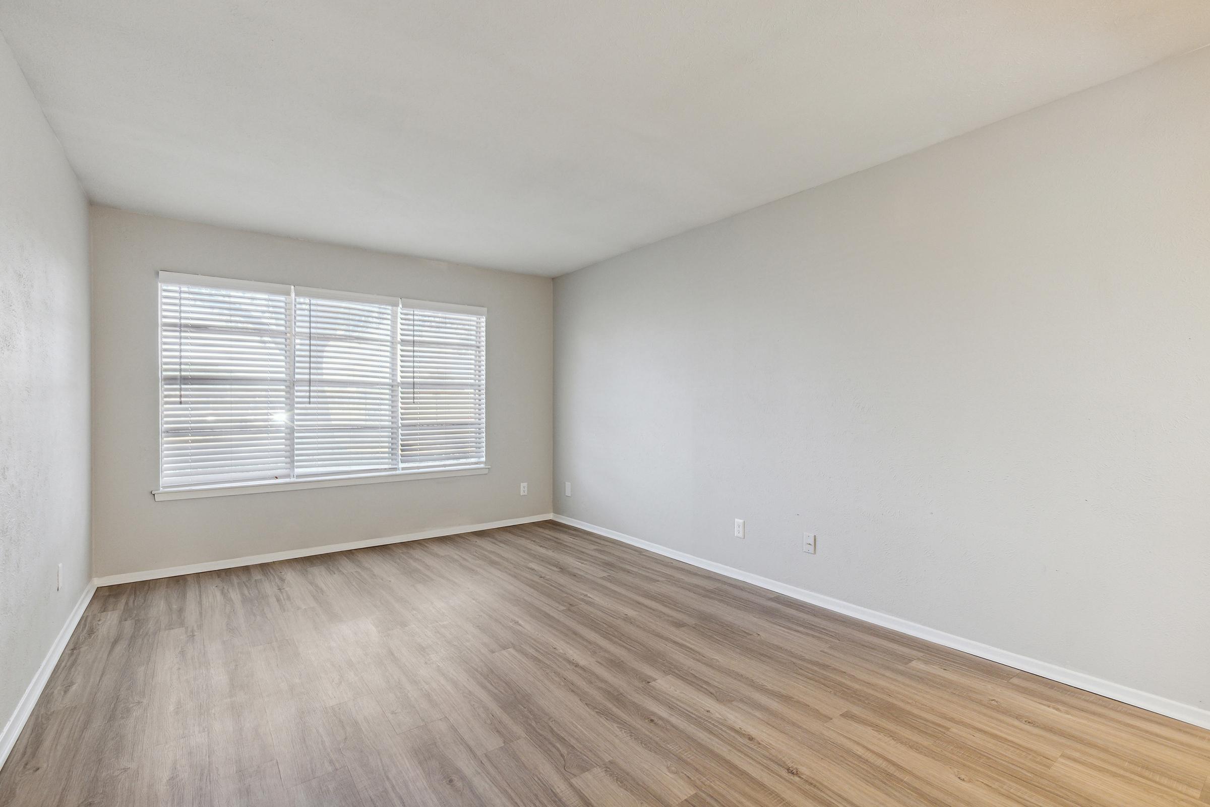 A clean, empty room featuring light-colored walls and wooden flooring. There are large windows with white blinds, allowing natural light to fill the space. The room has a simple, modern aesthetic with no furniture or decorations.