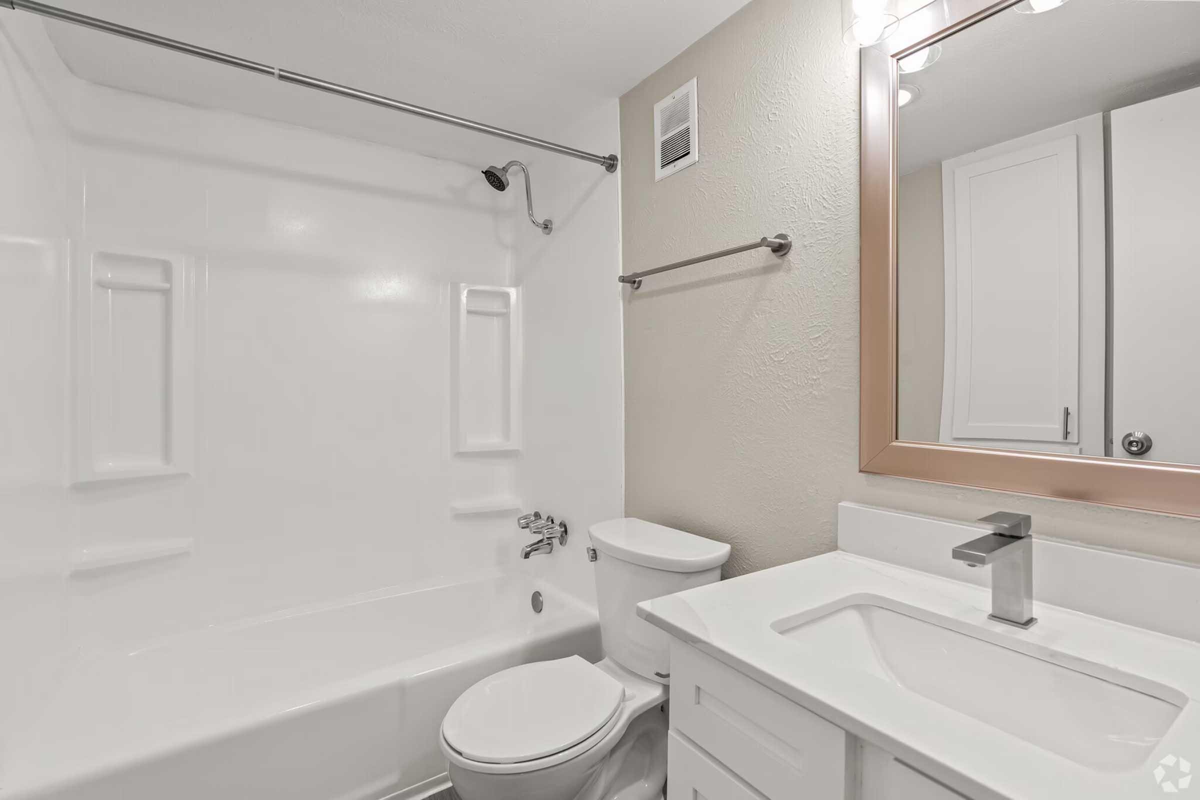 A modern bathroom featuring a white bathtub with a shower, a white toilet, and a sleek vanity with a sink and mirror. The walls are light-colored, and there are minimalistic fixtures, creating a clean and airy atmosphere.