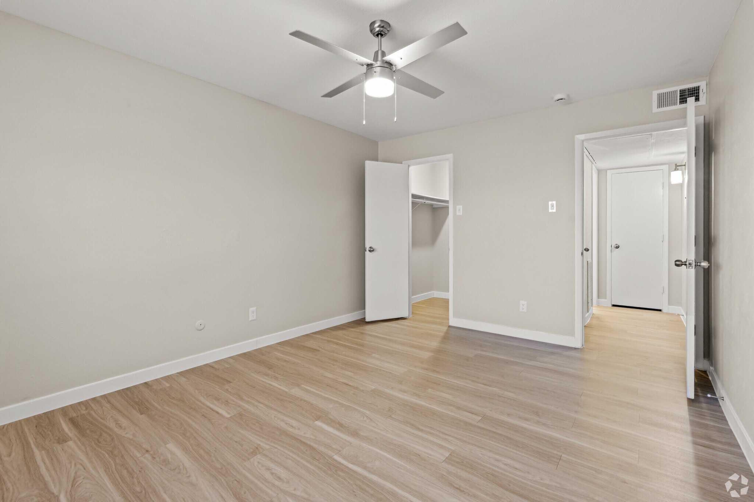 A minimalist room with light gray walls and wooden flooring. It features a ceiling fan, a closet with a white door, and an open doorway leading to a hallway. The space is well-lit and empty, ideal for personalization or furnishing.