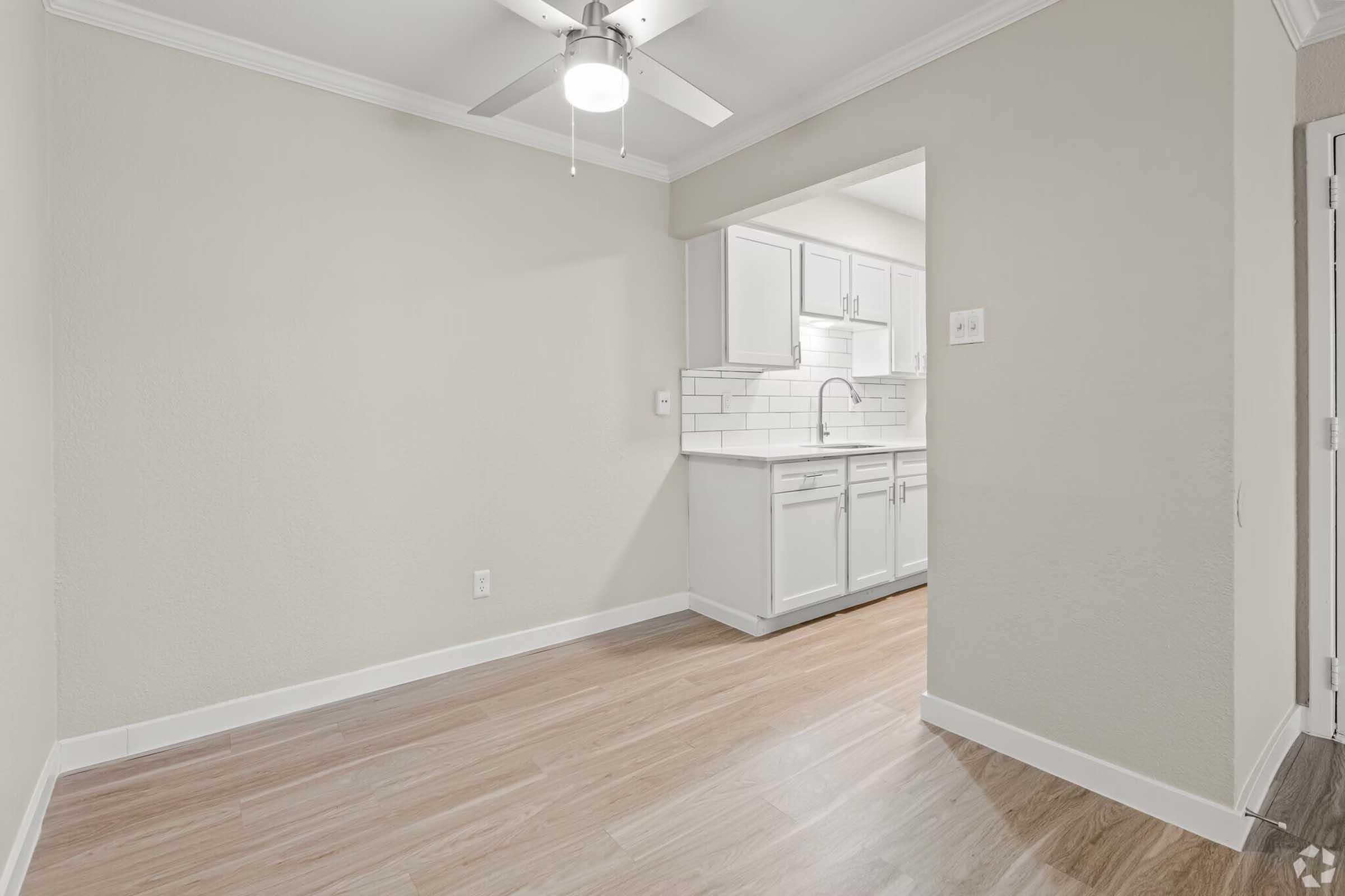 A well-lit interior space featuring light-colored walls, a ceiling fan, and wooden flooring. In the background, there is a kitchen area with white cabinetry and a tiled backsplash. The overall atmosphere is clean and modern, creating a welcoming environment.