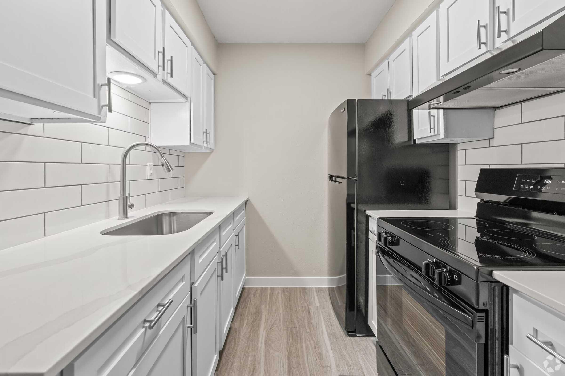 Modern kitchen featuring white cabinets, a stainless steel sink, and a black refrigerator. The countertop is light-colored, and the backsplash consists of white subway tiles. The kitchen includes an electric stove and oven, with wood-like flooring adding warmth to the space. The overall design is sleek and contemporary.