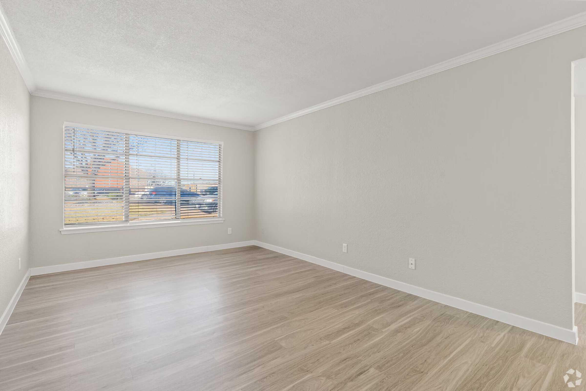 A spacious, empty room with light-colored walls and a large window. Natural light streams in through the window, illuminating the smooth, wooden floor. The room features white trim and a clean, minimalist design, creating a bright and airy atmosphere. Ideal for various uses, such as a living space or office.