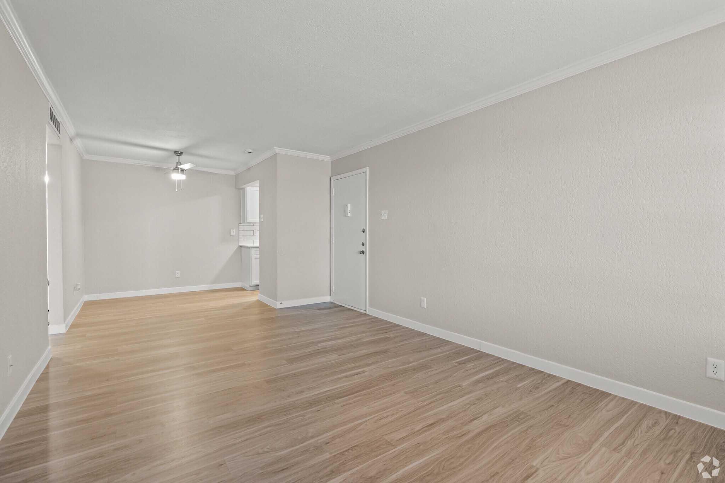 Spacious, empty living room featuring hardwood floors, light-colored walls, and a ceiling fan. A doorway leads to an entrance, and there is a glimpse of a kitchen area in the background. Natural light enhances the open feel of the space.