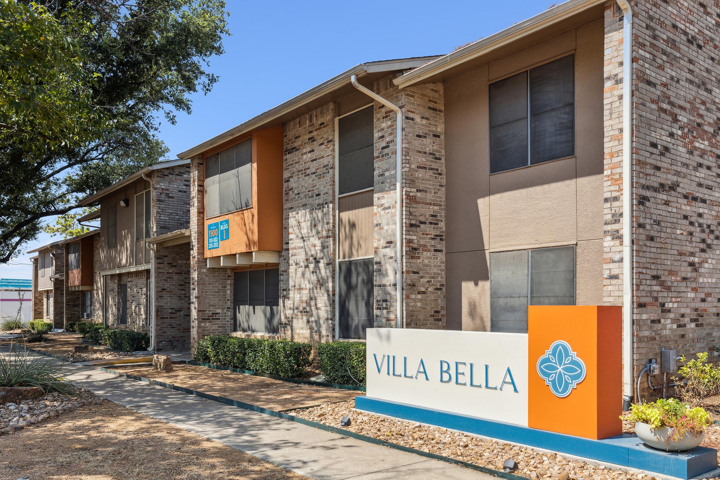 Exterior view of a residential apartment complex named "Villa Bella." The building features a mix of brick and stucco, with multiple windows and bushes lining the walkway. A prominent sign with the name "VILLA BELLA" is displayed in front of the building, set against a clear blue sky.