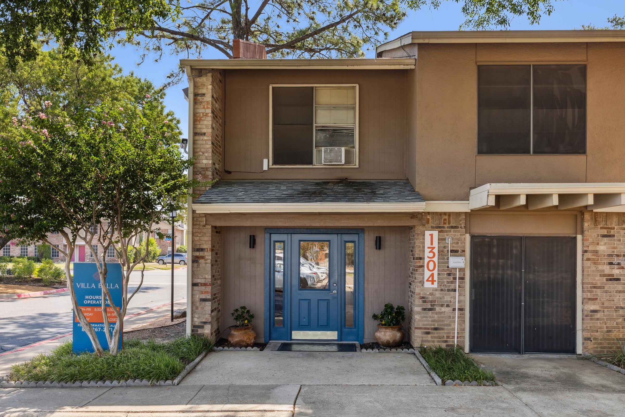 A residential building entrance featuring a blue front door with the number 1304 displayed. The facade includes brickwork and large windows, surrounded by greenery. A sign in the front yard displays “Vela Vista Apartments.” Clear skies and tree foliage add to the inviting atmosphere.