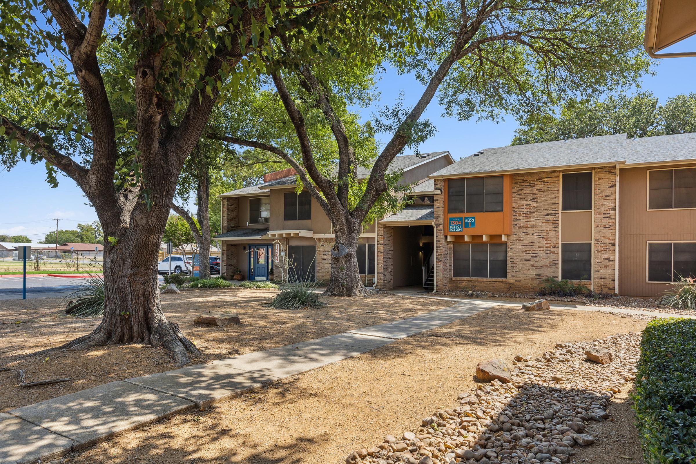 A residential building featuring two connected units with a stone and siding exterior. The pathway is lined with trees and shrubs, leading to the entrance. The area is sunny with clear blue skies, and a small rocky garden borders the path.