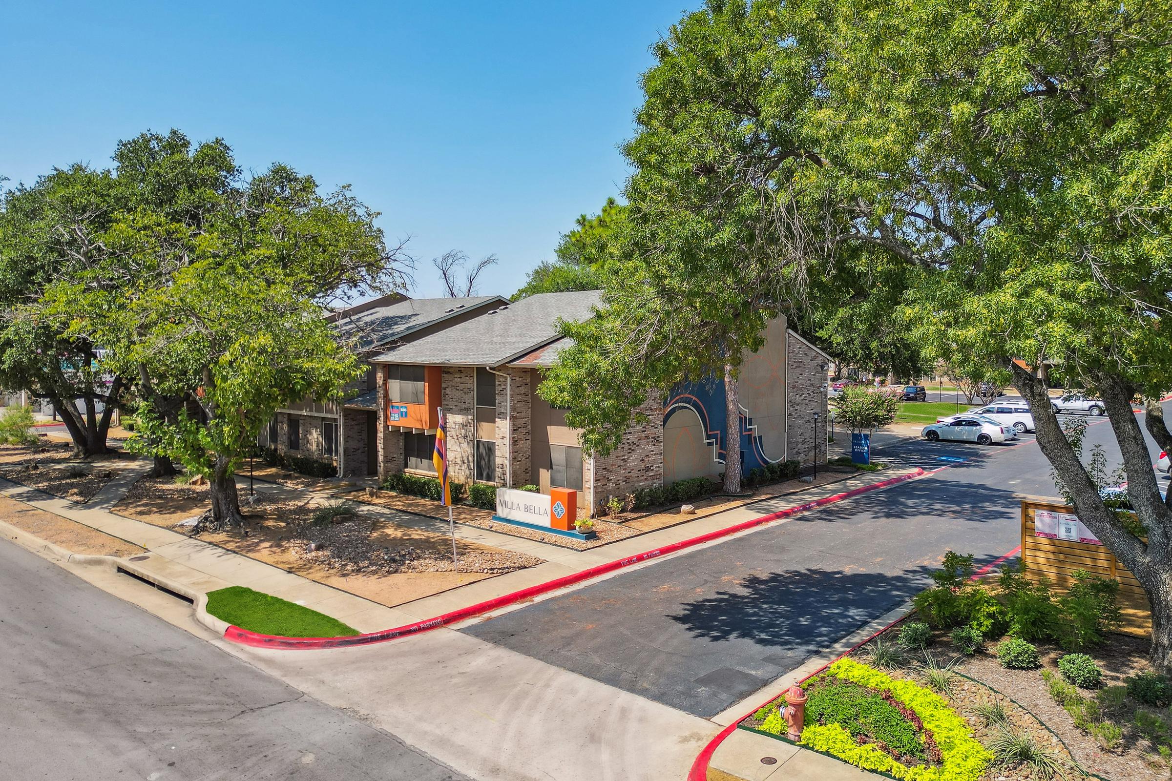 Aerial view of a residential building with a modern design, surrounded by trees and landscaped gardens. The entrance features signage, and there are parked cars along the driveway. The setting is sunny and clear, suggesting a warm, inviting atmosphere for residents.