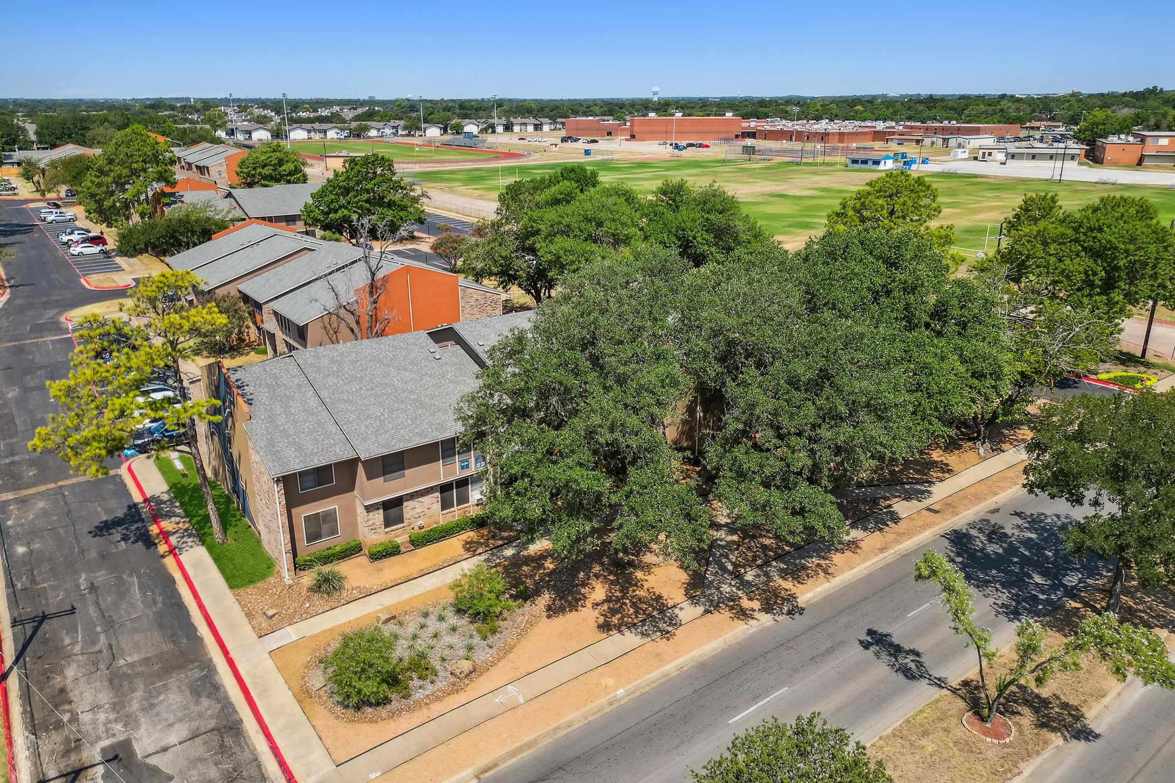 Aerial view of a neighborhood featuring residential buildings with trees, a parking area, and a grassy field or sports area in the background. The scene includes well-maintained landscaping and a clear sky, depicting a suburban setting.