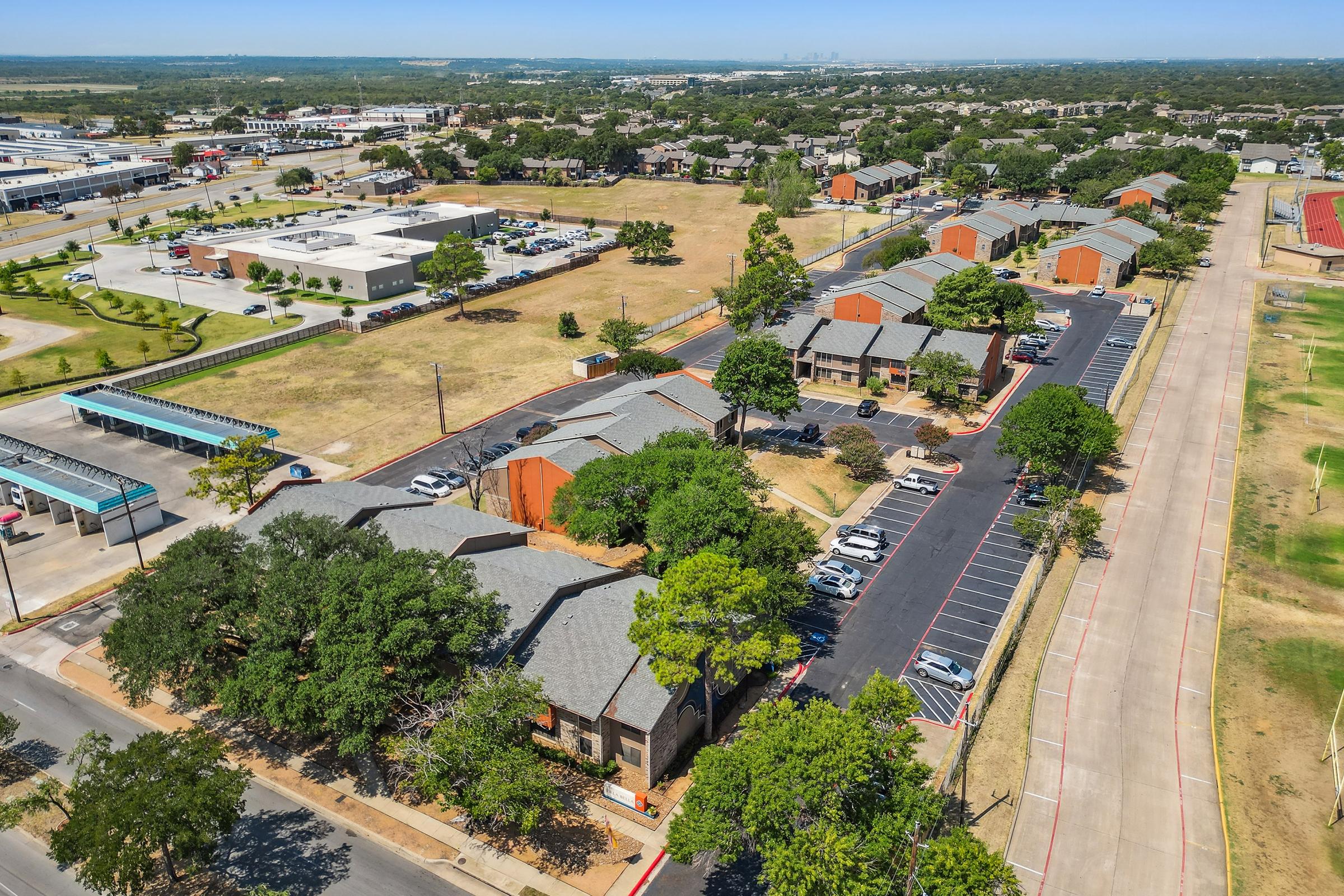 Aerial view of a residential area featuring multi-story buildings with parking lots. Lush green trees and landscaped spaces are visible, along with surrounding roads and a distant view of more urban development. The scene captures a sunny day with a clear sky.