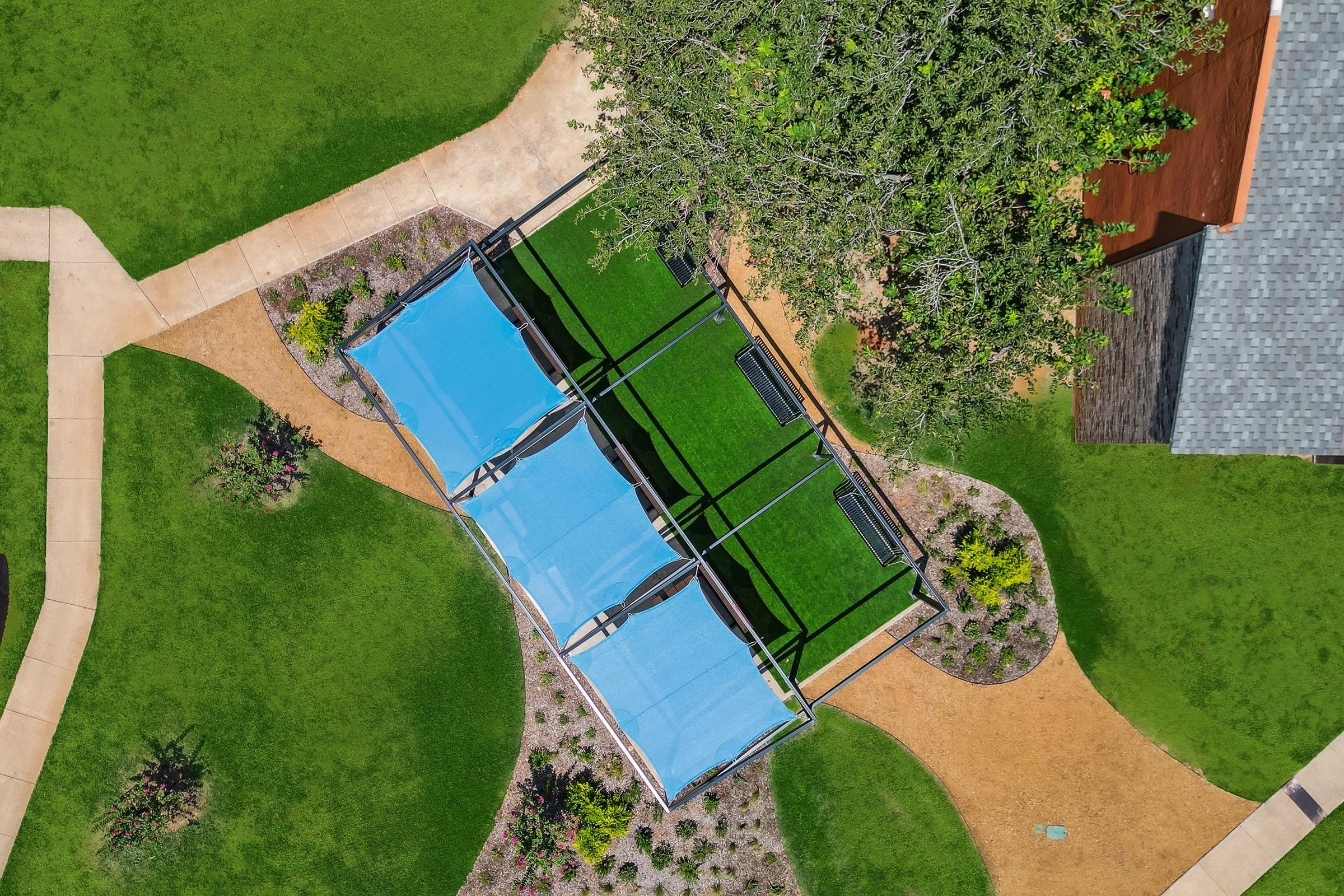 Aerial view of a park area featuring two blue shade structures over green turf, surrounded by neatly landscaped gardens and walking paths. The vibrant green grass contrasts with the tan pathways, and there is a well-maintained tree nearby, adding to the outdoor recreational environment.