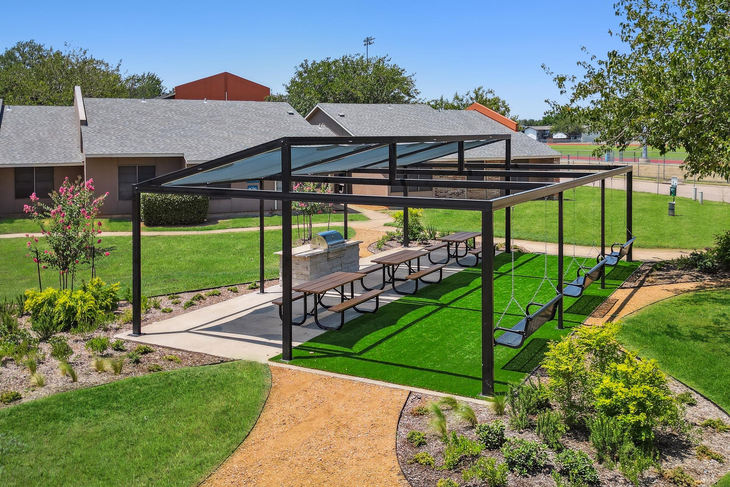 Outdoor pavilion featuring a gray steel frame with a glass roof, surrounded by green grass and landscaping. There are multiple picnic tables and swings, providing a recreational space. Nearby buildings and a baseball field can be seen in the background under a clear blue sky.