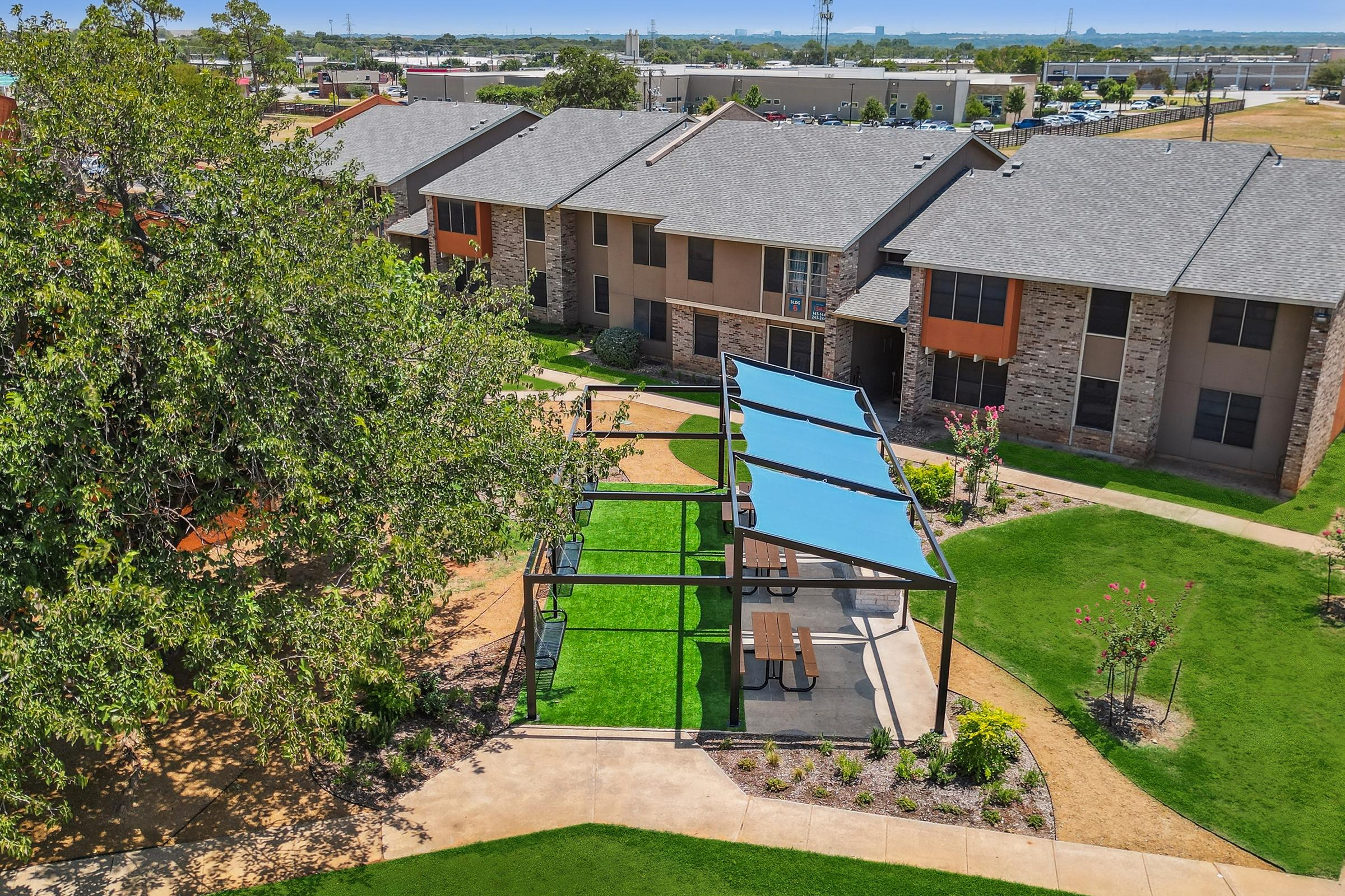 Aerial view of a residential complex showing two-story buildings with gray roofs. In the foreground, there's a landscaped area featuring green grass, a few picnic tables under blue canopies, and decorative shrubs, creating a communal outdoor space. The background includes more buildings and a distant view of industrial structures.