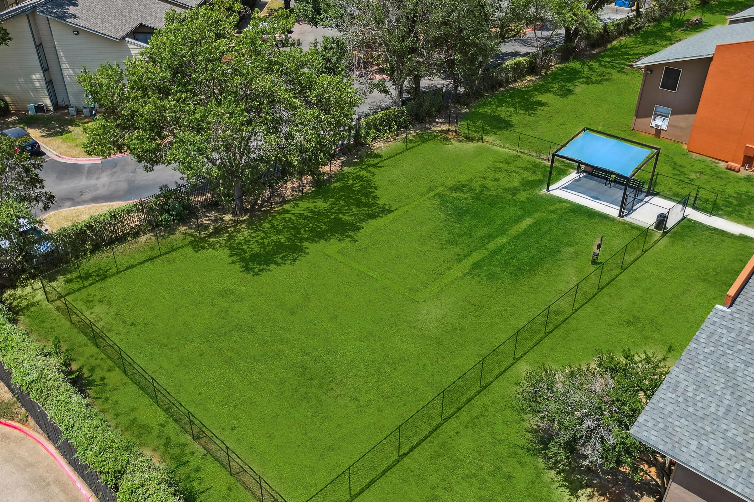 Aerial view of a green fenced yard with a small blue pavilion on one side. The yard is well-maintained, featuring lush grass and scattered trees. A pathway runs alongside the yard, with nearby buildings visible in the background.
